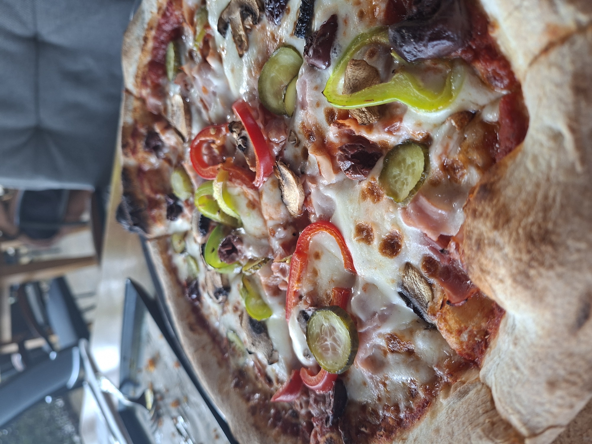 An overhead, close-up, slightly tilted shot shows a savory pizza with a thick, golden-brown crust. The pizza is generously topped with melted white cheese, red sauce, sliced mushrooms, green and red bell peppers, pickles, and black olives. The pizza rests on a wooden table, with a corner of a dark plate and a fork visible on the left. In the background, a blurred scene of chairs and a table suggests a dining establishment.