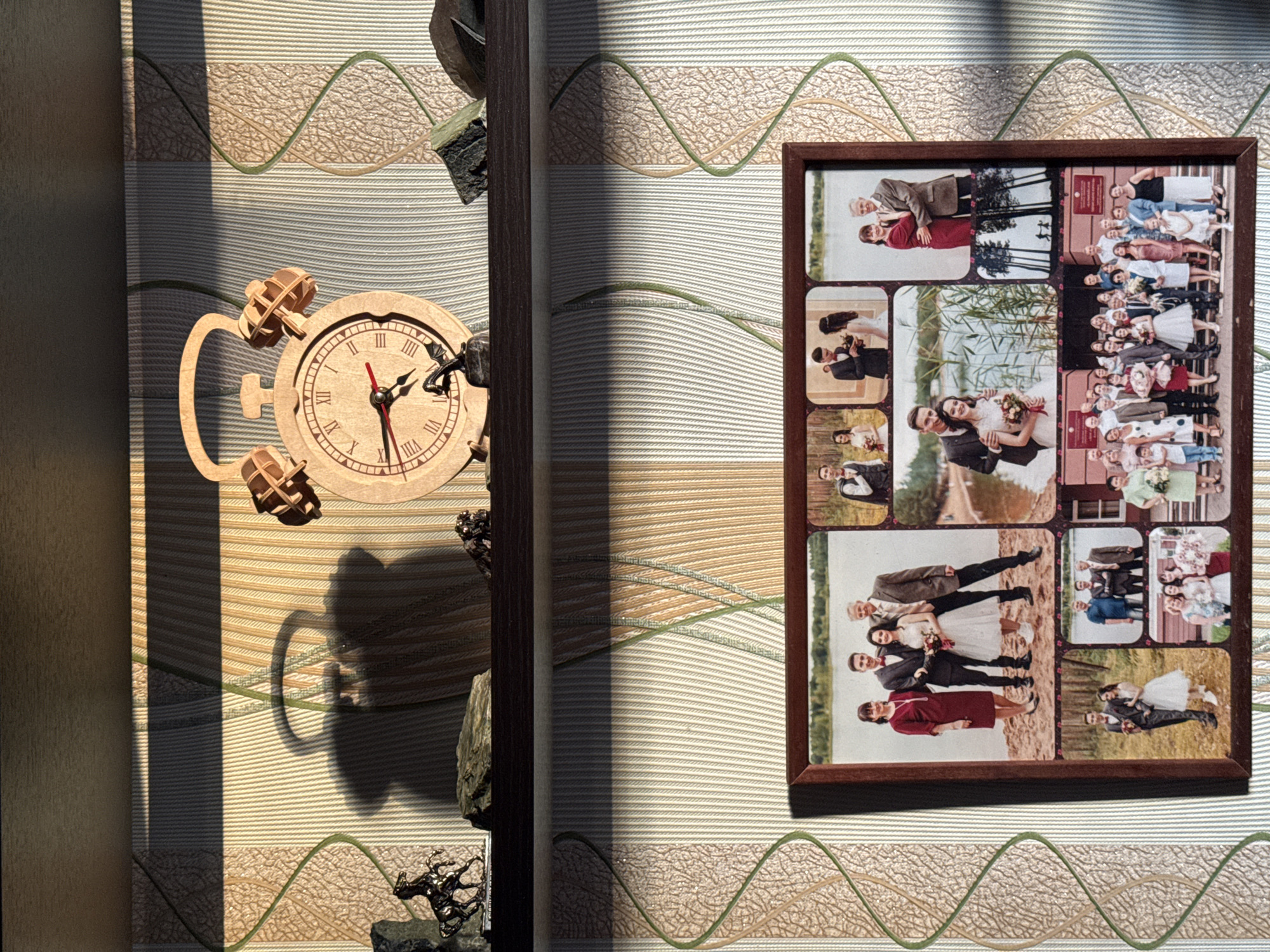 The image shows a top-down view of a textured surface, possibly a wall covering, with a light and shadow pattern. On the left, there's a wooden clock with the hands showing approximately 2:52. Beside it, there are wooden gears and figurines on a piece of stone. To the right of the clock is a dark vertical structural element, likely part of a frame or wall feature. On the right side, there's a rectangular framed collage of wedding photos. The location of the photo is possibly in Izhevsk, Russia. There is no other evidence to verify the exact location in that city.