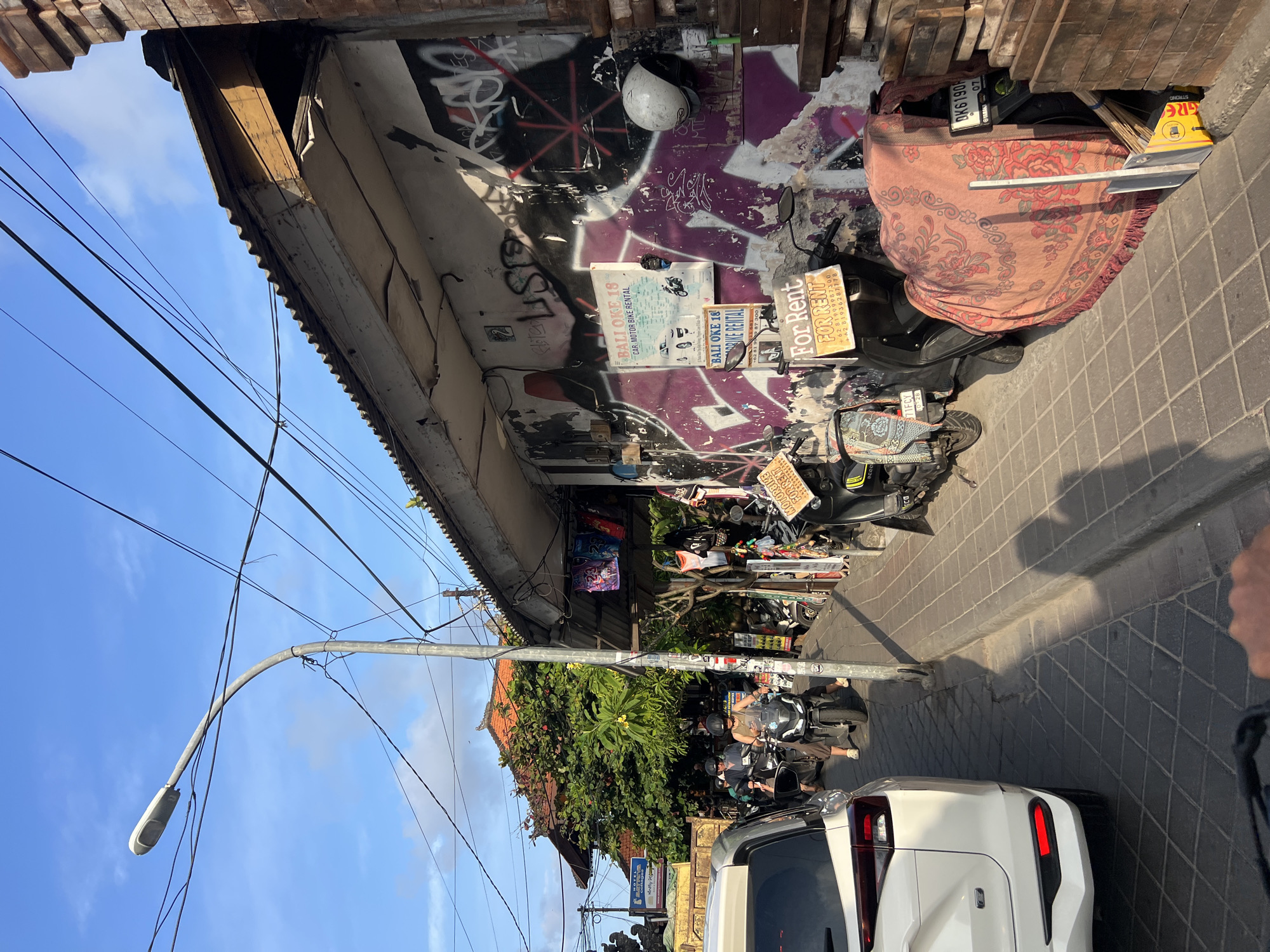 The image captures a street scene in Kuta, Indonesia, likely a commercial area with shops and rental services. The foreground shows the back of a white SUV, and to its right, a sidewalk paved with interlocking gray pavers. A street lamp with a curved pole stands on the sidewalk, surrounded by a tangle of electrical wires against a bright blue sky with scattered clouds.
The buildings lining the street have weathered facades covered in graffiti. One building has a corrugated metal roof and exposed wooden beams. Several motorcycles are parked along the sidewalk, some with "For Rent" signs attached. One scooter is draped with a patterned textile. There are also signs advertising "Bali Oke 18" for car and motorbike rental.
In the middle ground, several people are visible on motorcycles, navigating the street. Some are wearing helmets. In the background, more buildings with traditional tiled roofs and lush green trees are visible. The overall impression is of a busy, tropical urban environment.