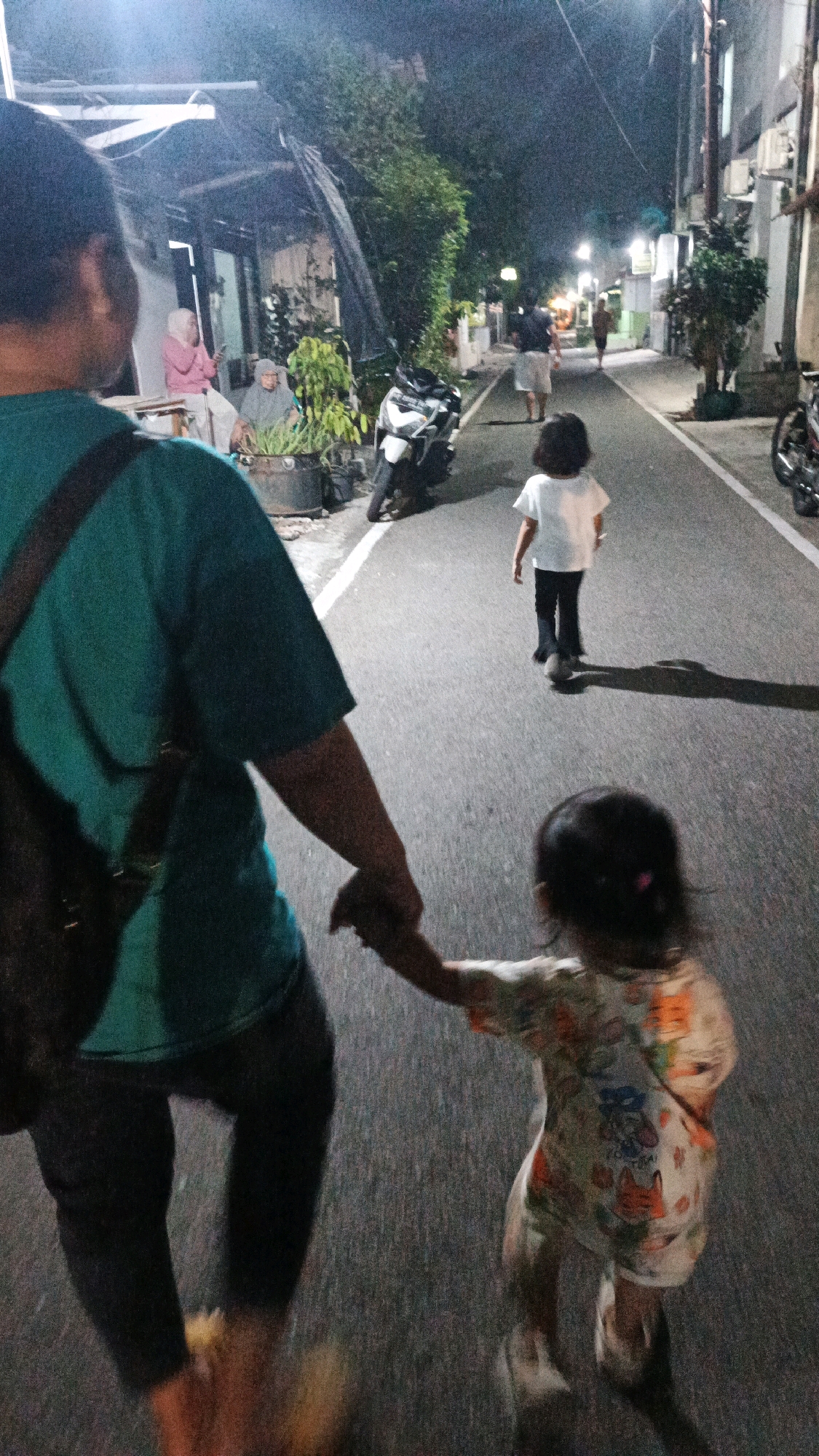 This is a night-time, street-level, medium-shot photograph taken from a first-person perspective looking down a paved street in Surakarta, Indonesia. In the foreground, the back of an adult, wearing a teal t-shirt and dark pants, is visible, holding the hand of a young child. The child, also viewed from the back, is wearing a light-colored, patterned shirt and dark pants, and is walking away from the camera. Further down the street, another child, wearing a white t-shirt and dark pants, is also walking away. Several buildings with illuminated windows line the right side of the street. On the left, there are some plants and a parked motorcycle. The overall impression is of a casual evening walk.