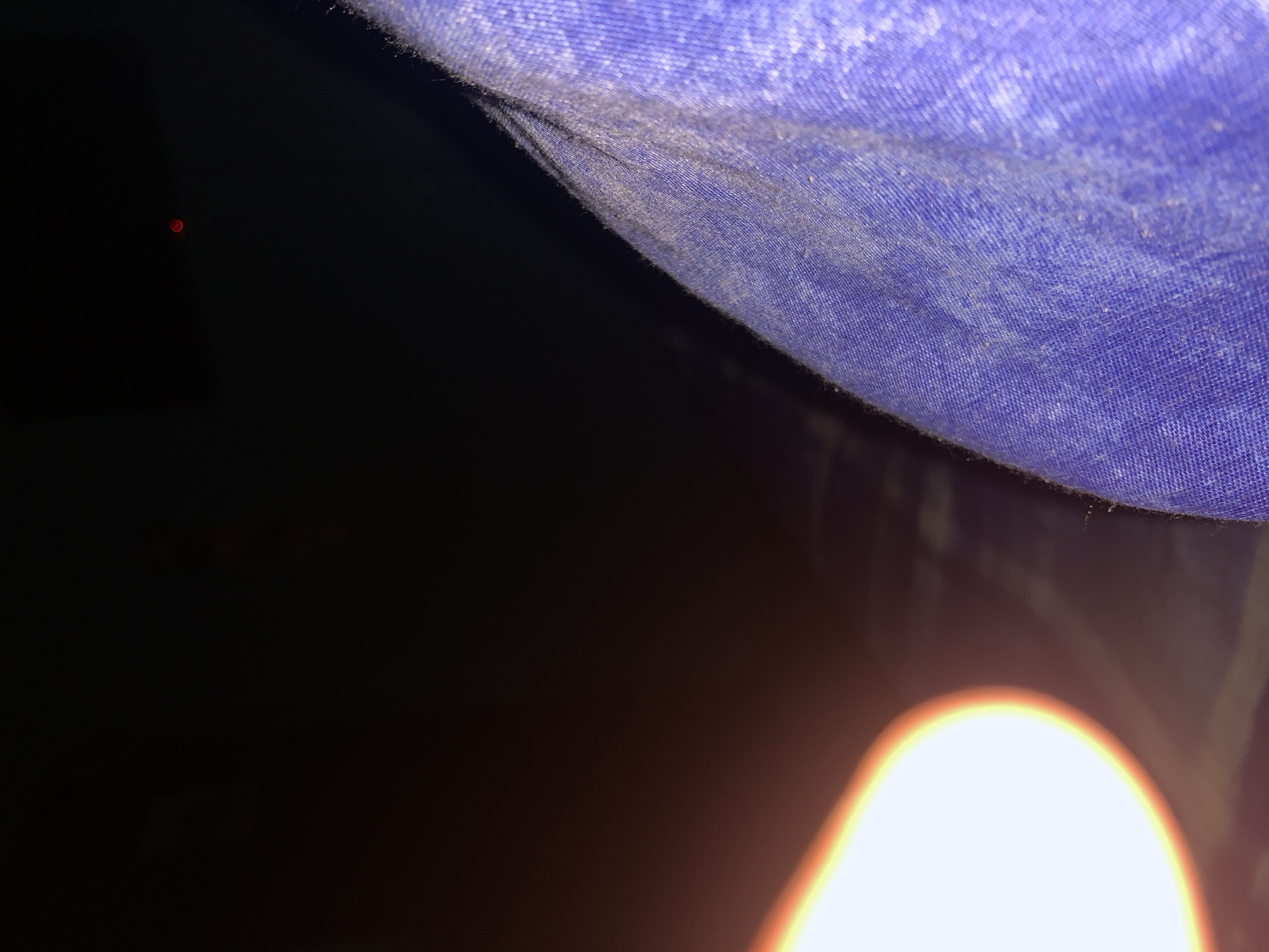 The image presents a close-up, angled view inside a dark, confined space. The upper and right portions of the frame are dominated by a textured, purplish-blue fabric, likely made of synthetic material such as nylon or canvas. This fabric exhibits visible dust and dirt particles, suggesting wear or exposure to an outdoor environment, possibly indicating a tent or similar shelter. The weave of the material is clearly discernible.
The majority of the lower and left areas of the image are enveloped in deep shadow, almost completely black, signifying extremely low light conditions. In the bottom right corner, a bright, out-of-focus light source emits an intense orange-yellow glow, illuminating a portion of the darkness and subtly reflecting off the edge of the blue fabric. This light appears to be very close to the camera, possibly from a flashlight or mobile device. A tiny, faint red light dot is visible in the distant dark background towards the upper left corner.
The scene conveys being within a sheltered, dark enclosure, likely at night or in an unlit interior. The material and dirt suggest an outdoor context or a rugged setting in Santa Ana, Venezuela. There are no visible people or text.