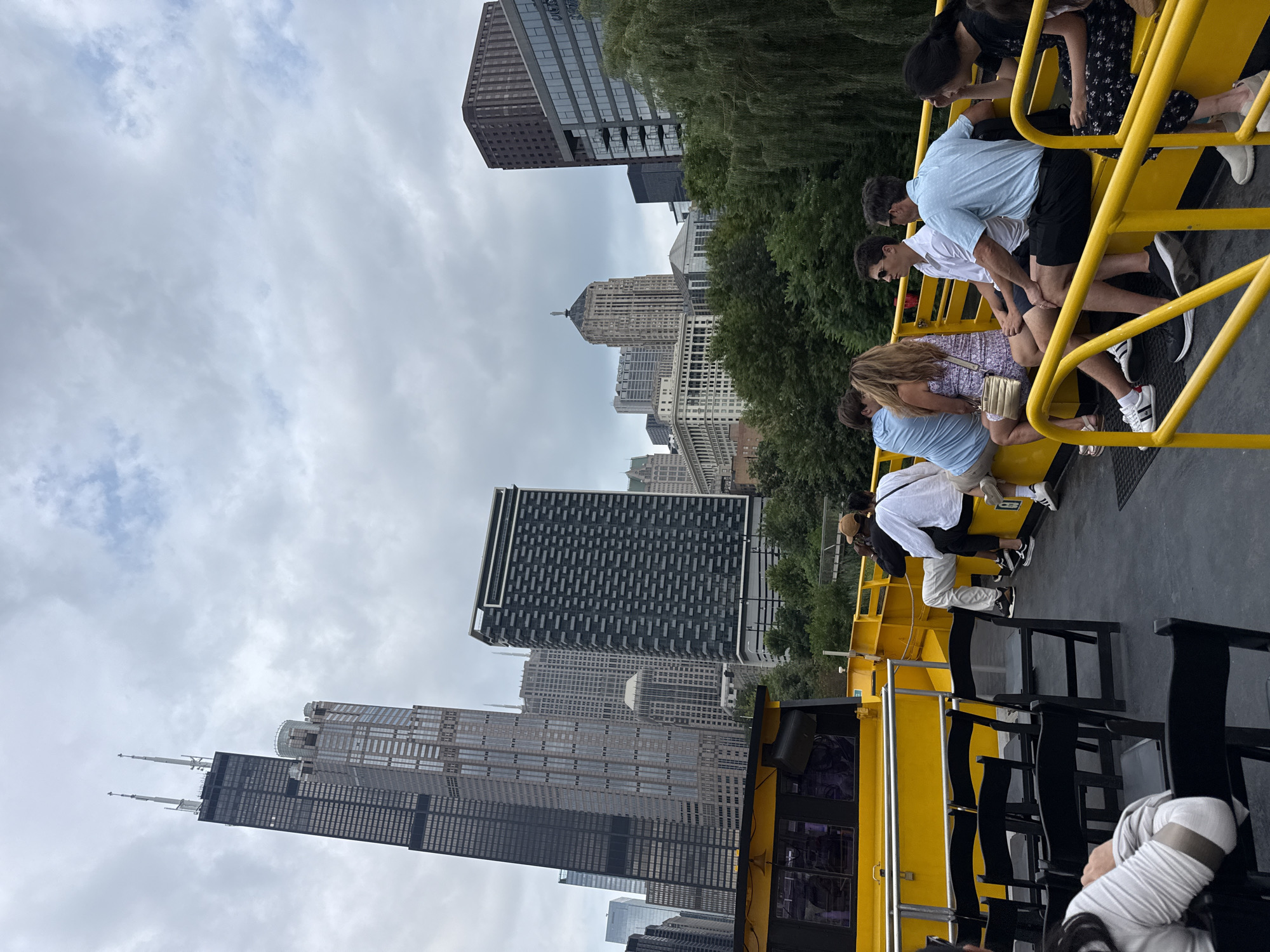 In Chicago, Illinois, on a cloudy day, a group of people are seated on a bright yellow tour boat. The boat's deck is visible, with black folding chairs arranged in the foreground. The people on the boat are dressed casually, with some facing away from the camera and others looking towards the cityscape. In the background, a dense cluster of skyscrapers dominates the skyline. Prominent among these is the Willis Tower, formerly known as the Sears Tower, a dark, rectangular skyscraper with distinctive antennas. Other modern and older architectural styles of buildings are also visible, interspersed with lush green trees. The overall impression is of a bustling urban environment viewed from a leisurely tour.