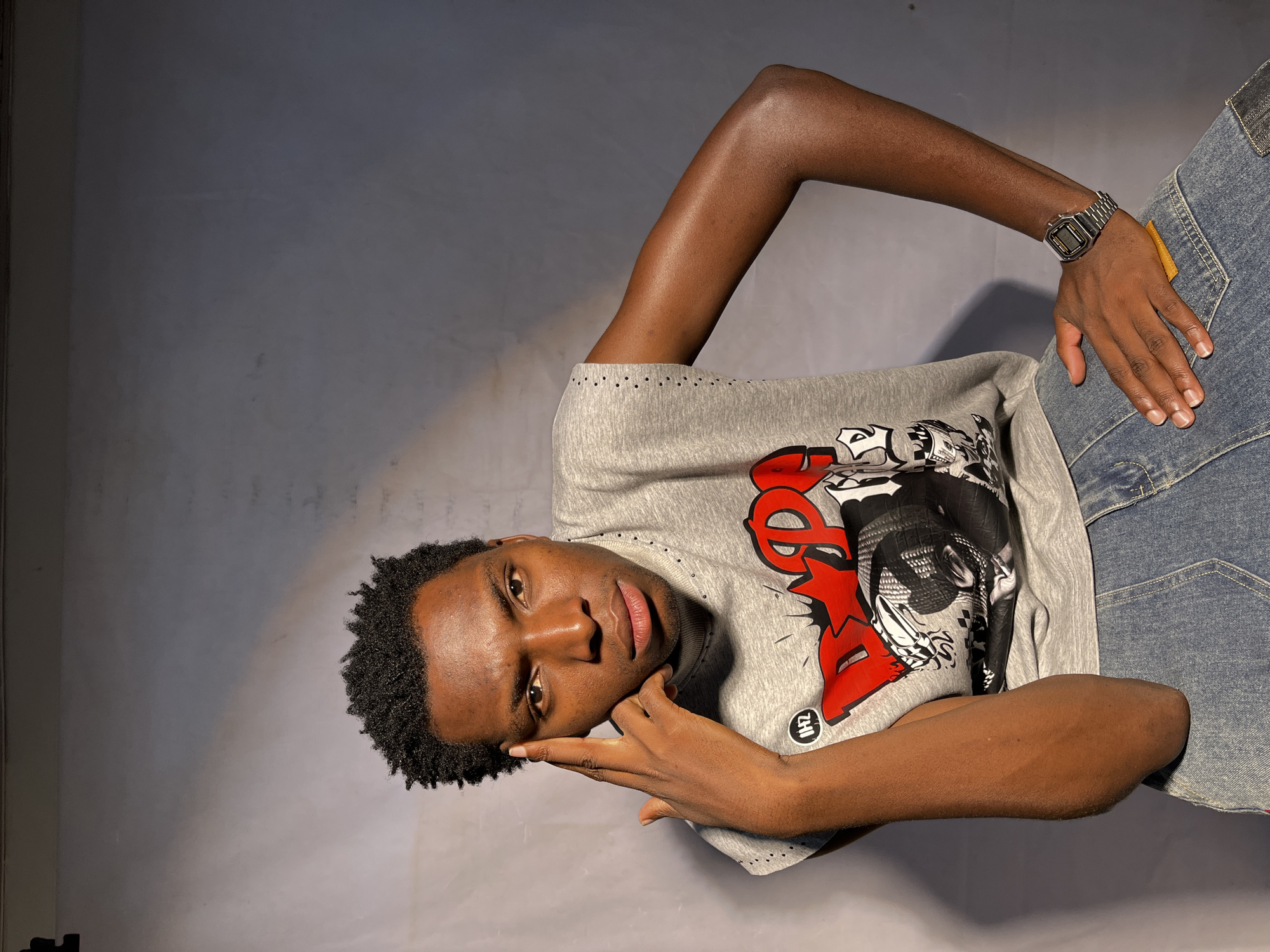 A young Black male with short, dark, curly hair sits on a low stool in a studio setting. He is wearing a grey sleeveless t-shirt with a graphic print and blue denim jeans. His left hand is placed on his forehead, with his fingers touching his temple. His right hand rests on his thigh. A silver watch is visible on his left wrist. The background is a plain, grey wall. This image was taken in Barakin Akawo, Nigeria.