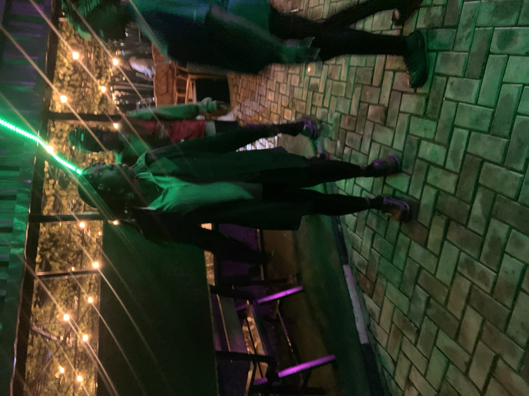 In Gura Top, Nigeria, three individuals are captured in a dimly lit outdoor setting, likely an establishment with a covered seating area. A green light strip runs diagonally across the top of the image, illuminating the scene with a vibrant glow. The ground is paved with interlocking rectangular bricks, some of which appear wet. In the background, a series of warm-toned lights twinkle among foliage, suggesting string lights in trees or plants.
The person in the foreground, facing left, is wearing a dark trench coat over a light-colored shirt. They are looking away from the camera, and their left leg is forward, indicating movement. To their right, a person in a red vest over a green garment is also in motion, facing away from the camera and towards the right. Further to the right and slightly behind, a third person, mostly obscured by shadow, is visible. This individual is wearing dark clothing and their right leg is forward.
In the middle ground, a dark wooden bench with purple accents is partially visible. The overall atmosphere is suggestive of a relaxed evening gathering at a venue in Gura Top.