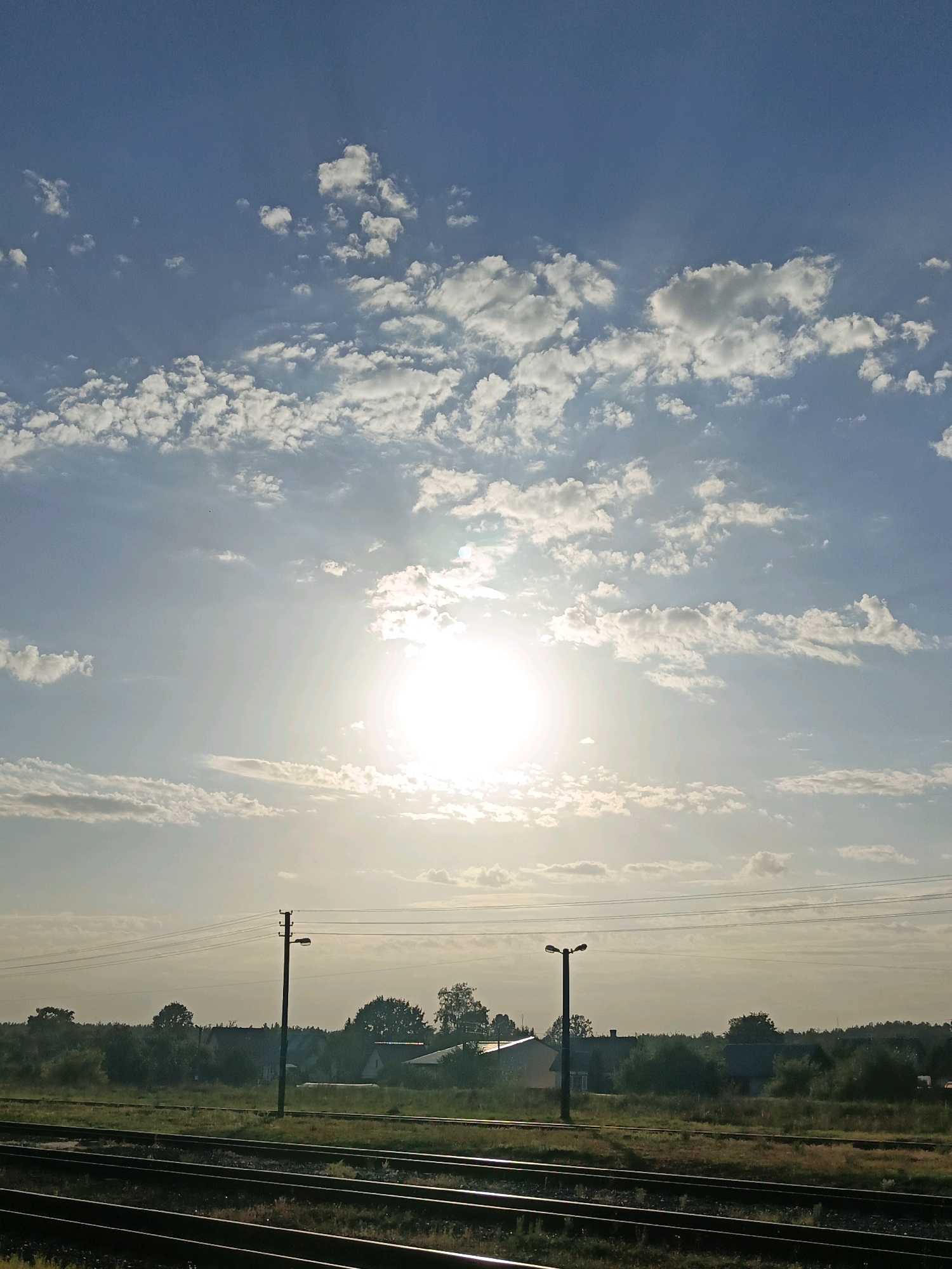 The image depicts a rural landscape with a sky dominated by a bright sun partially obscured by scattered clouds. In the foreground, multiple sets of railroad tracks are visible, running horizontally across the frame. Beyond the tracks, a treeline separates the railway from a small village, identifiable by the rooftops of several houses and buildings. Two lampposts stand tall in the middle ground, with power lines stretching between them and across the scene. The environment suggests an outdoor setting in a semi-urban or rural area, possibly in the vicinity of Парэчча, Belarus, given the presence of infrastructure like train tracks and village housing amidst natural vegetation. The lighting indicates it is daytime, with the sun casting a bright glow. There are no people or animals visible in the image.