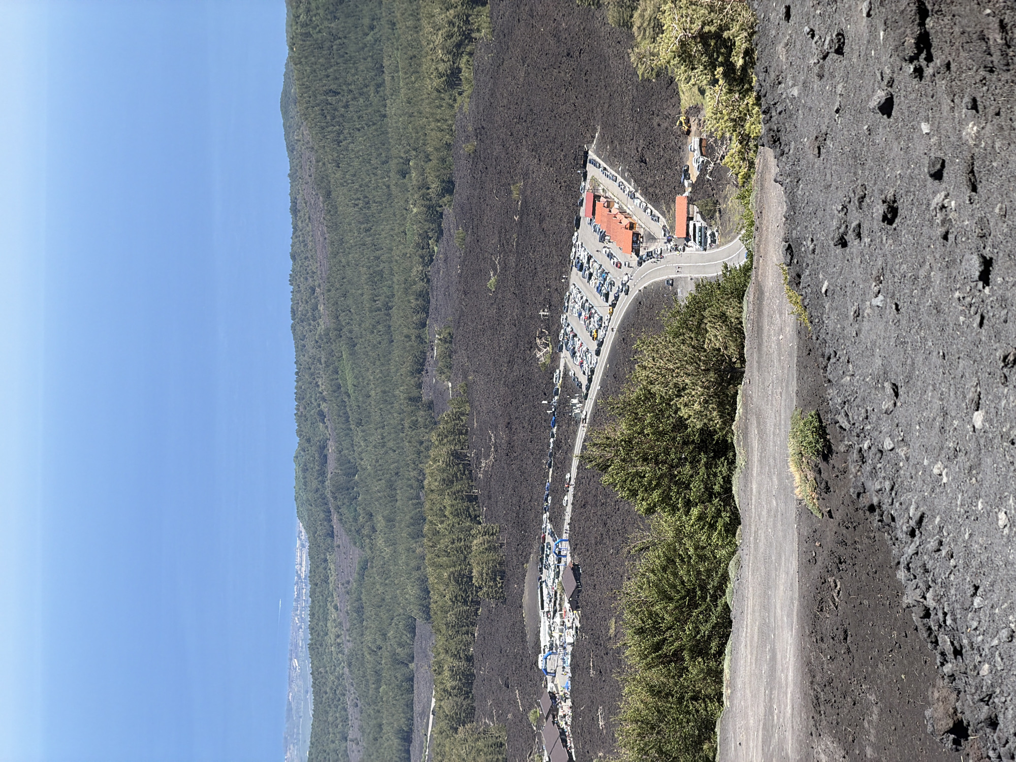 A high-angle, full shot captures the rugged, dark terrain of Mount Etna near Linguaglossa, Italy. The foreground features a steep, dark slope covered in volcanic rock and ash, with sparse patches of green vegetation. In the mid-ground, a paved road winds through a vast expanse of solidified lava flow, marked by dark, uneven surfaces. Along the road, several small buildings with orange-tiled roofs are visible, likely serving as visitor facilities. A large parking lot is filled with numerous cars, and a few tour buses are parked nearby. Beyond the lava field, a dense forest of green trees stretches across the landscape. In the background, the vast expanse of the blue Mediterranean Sea meets the horizon under a clear sky. The scene depicts a popular tourist area on a volcanic mountain, with infrastructure designed to accommodate visitors exploring the unique geological environment.