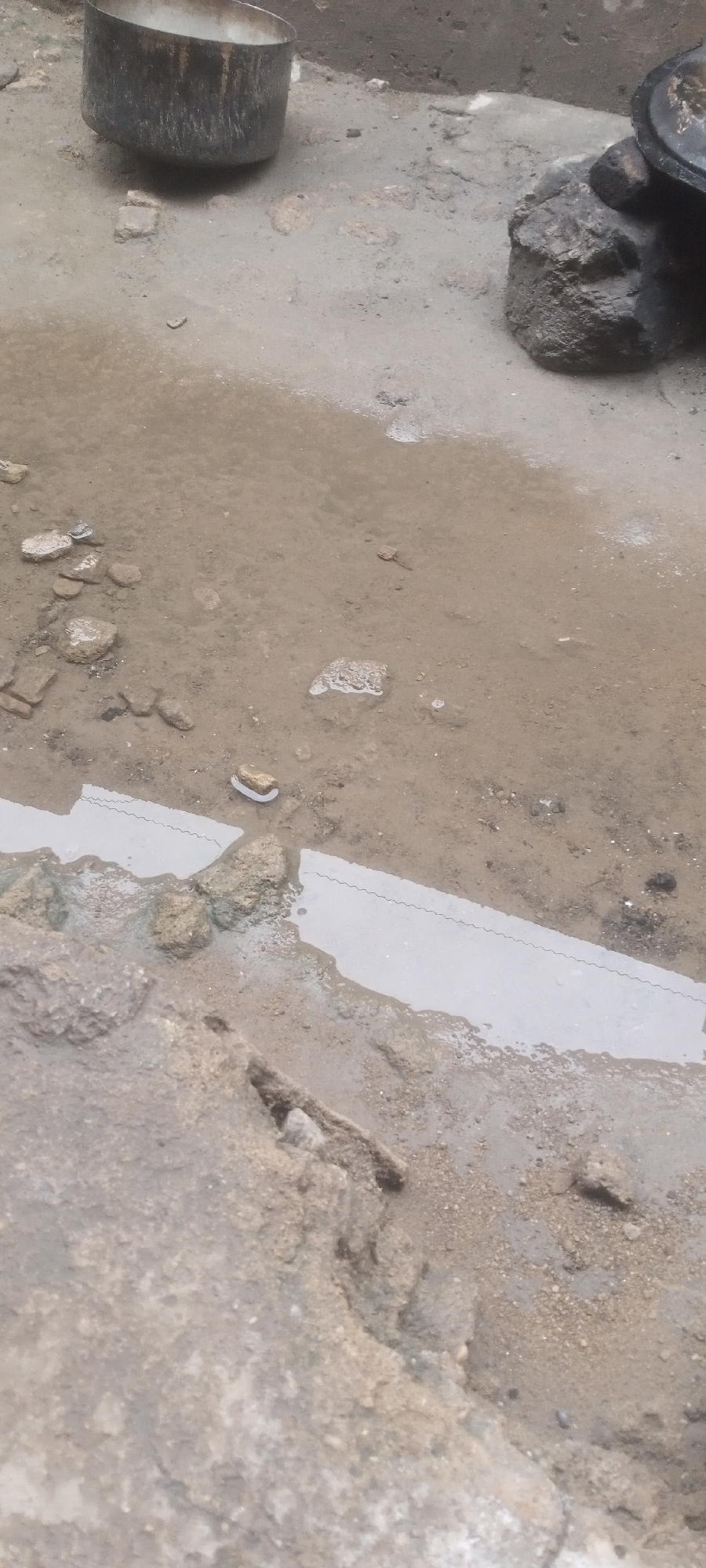The scene is a puddle in a muddy area in Sabon Tasha, Nigeria. A metal pot sits in the background, with rocks nearby. The ground is uneven and wet. Part of a concrete structure is visible in the foreground.