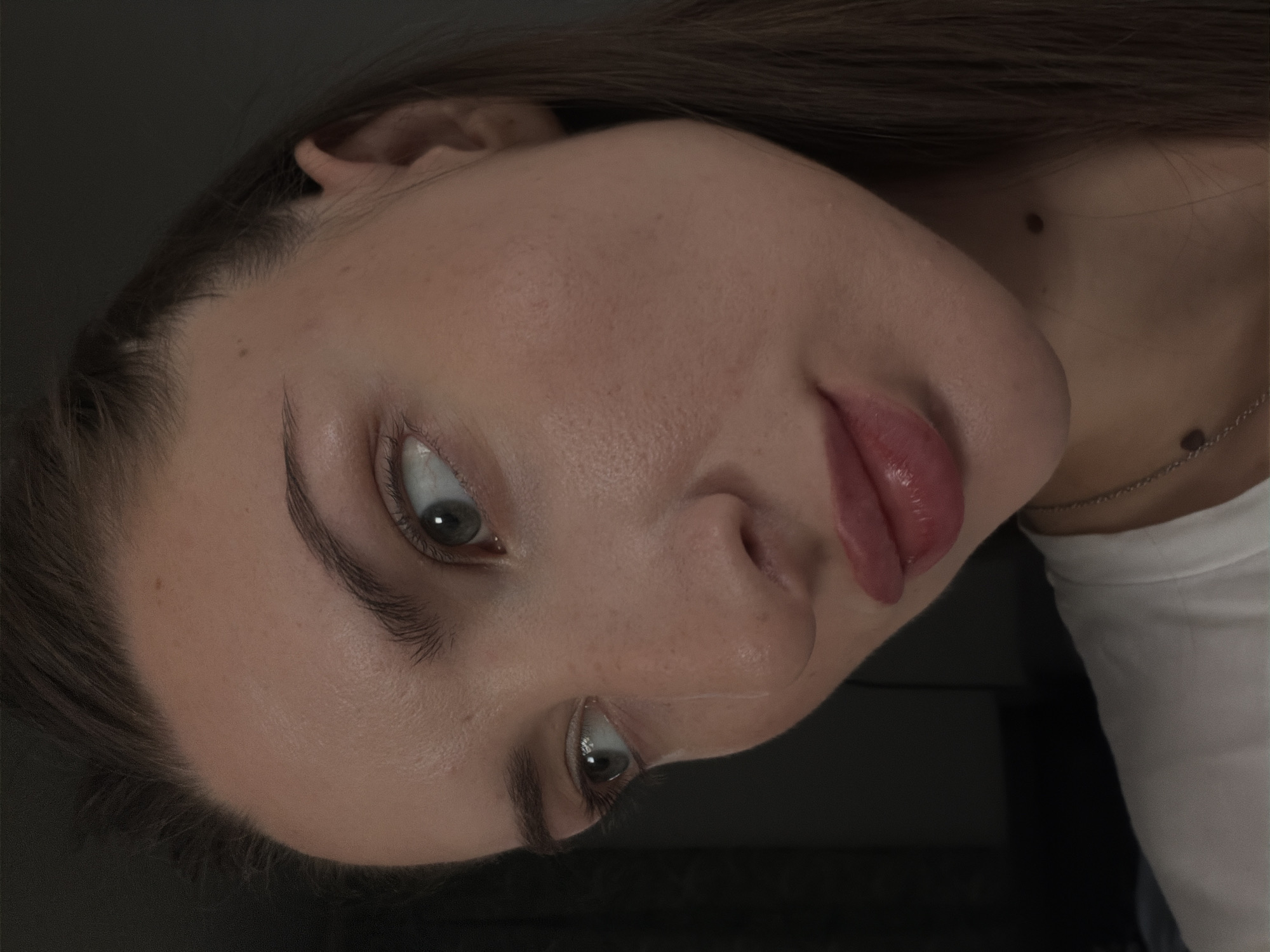 The image is a close-up, high-angle, eye-level shot of a young Caucasian woman's face, captured in a dimly lit indoor setting. The woman has fair skin with some visible pores and a few small moles on her face and neck. Her left eye is looking towards the left side of the frame, and a tear track is visible on her cheek below it. Her eyebrows are dark and well-defined. Her lips are full and appear to be wearing a glossy pink or coral-toned lipstick. Her hair is dark brown and pulled back partially. She is wearing a white t-shirt and a delicate silver necklace. The background is dark and out of focus, suggesting a plain wall or backdrop. The image quality is generally good, with some noise visible. The provided information about the location in Sharm El Sheikh, Egypt, is not supported by any visual cues in the image itself.