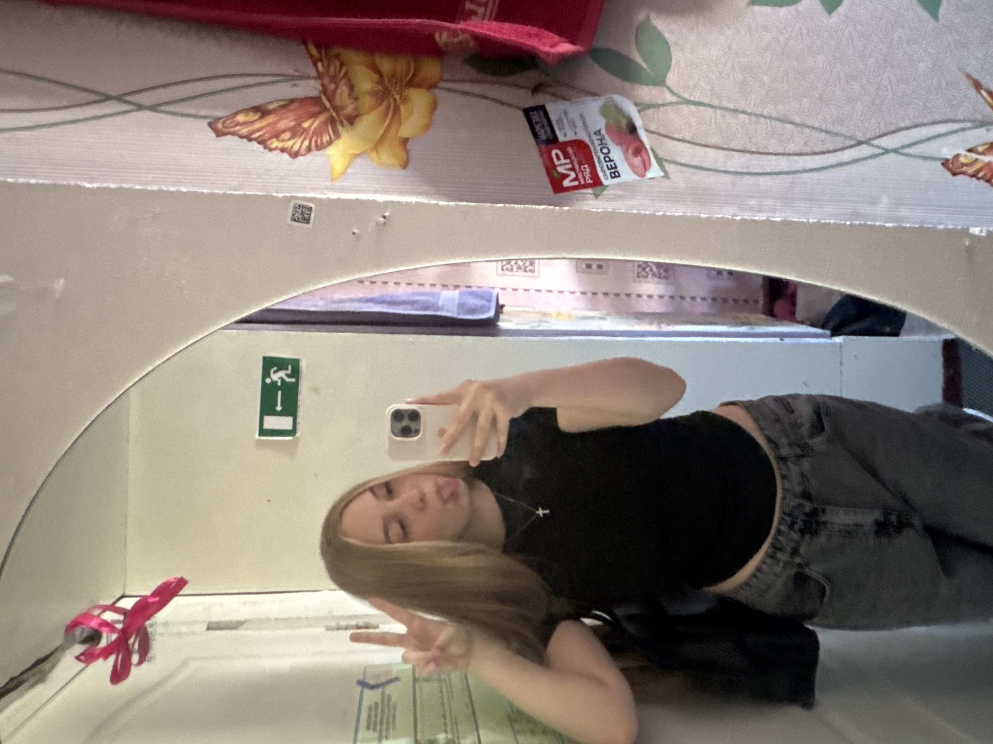 The image is a mirror selfie, rotated 90 degrees clockwise, depicting a young woman. She is positioned horizontally, appearing to lie on her side, in front of a mirror with a subtly arched top. The woman has long, straight light brown hair and is wearing a black short-sleeved top and dark grey jeans. She holds a light-colored smartphone in her right hand to take the picture, while her left hand forms a peace sign. Her expression is playful, with her tongue slightly extended. She wears a small cross pendant necklace.
Reflected in the mirror, behind the woman, is a green and white emergency exit sign featuring a running person pictogram and an arrow pointing to the right. A small pink ribbon or plastic decoration is visible near the mirror's edge. A black bag strap is seen near her lower back.
Above the mirror, showing the actual room decor behind the camera, is wallpaper adorned with a pattern of yellow and orange butterflies or flowers and green vines on a light background. A small, rectangular white and red sachet is resting on this patterned surface. Visible Cyrillic text on the sachet includes "MP", "РИД", and "ВЕРОНА", along with smaller text "САХАРОЗАМЕНИТЕЛЬ" (sugar substitute) and "ЯГОДНЫЙ" (berry), indicating it's a berry-flavored product. A tiny, pixelated QR code is also present on the wall above the mirror. A red object, possibly fabric, is at the top edge of the frame.
The scene suggests an indoor setting, likely in Perm, Russia, as evidenced by the Cyrillic text. The overall mood is casual and lighthearted.