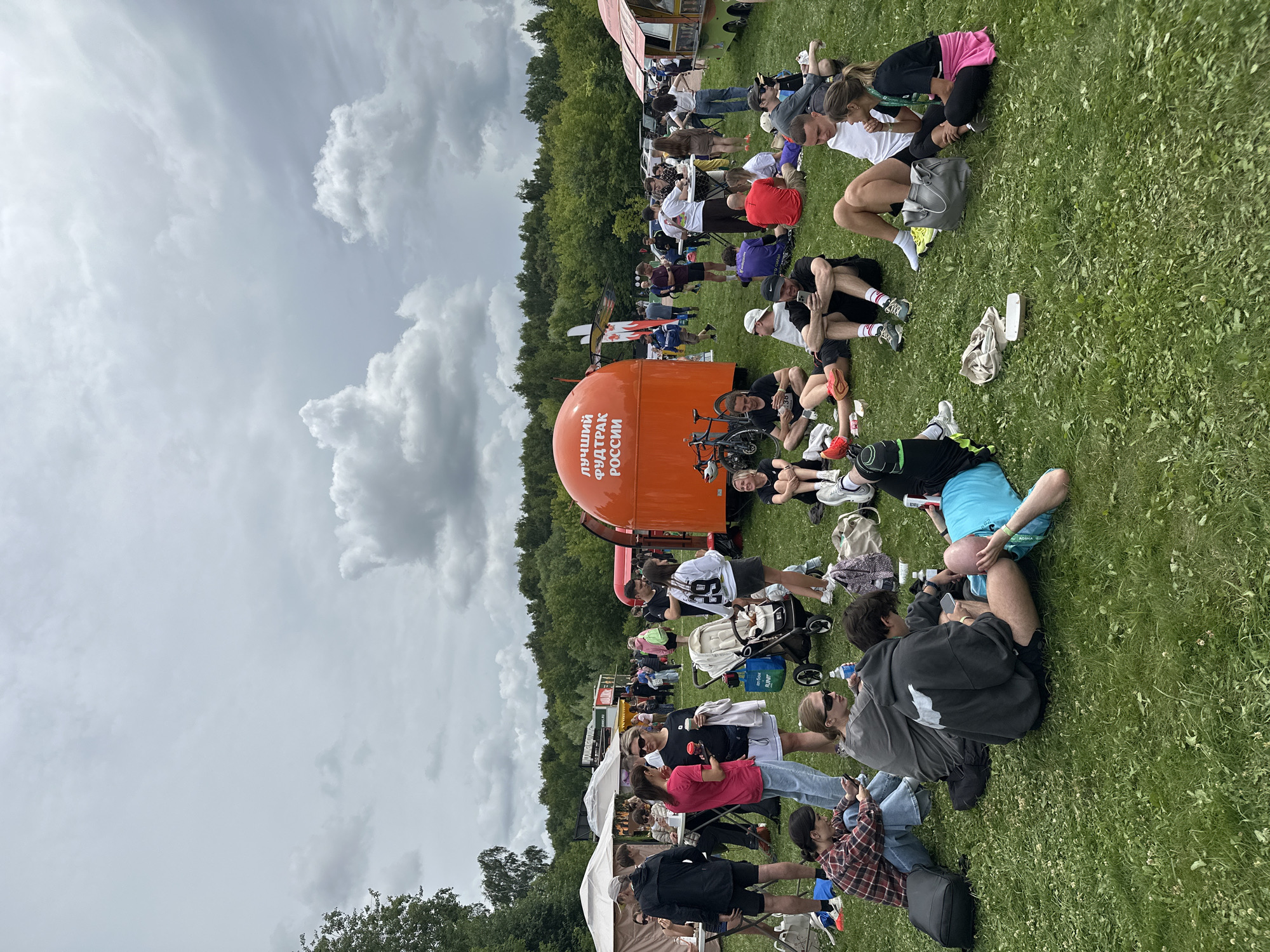 The image depicts an outdoor event with many people gathered on a grassy field. In the foreground, several individuals are sitting and lying on the grass, some interacting with their phones, others chatting. A stroller is visible, suggesting families are present. In the middle ground, a large orange food truck with Russian text is parked, alongside other food stalls and tents. People are standing and walking around these stalls, indicating a food festival or similar gathering. The background features a line of trees, typical of a park or outdoor venue. The sky is overcast with dramatic clouds. The event appears to be taking place in a park-like setting within the city of Moscow, Russia, likely during a summer day given the clothing choices of the attendees.