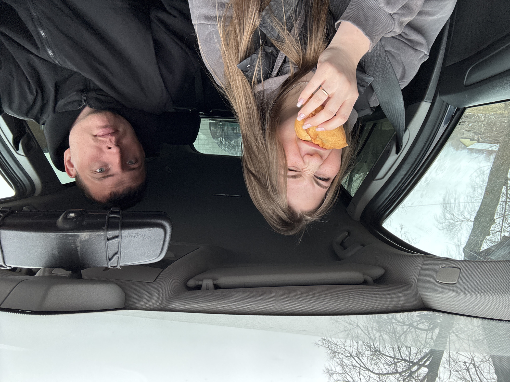 From inside a car, a selfie shows a man and a woman. The man on the left has short brown hair and wears a black jacket. The woman on the right has long brown hair and wears a gray hoodie. She is holding a pastry near her mouth and is making a funny face. The car's rearview mirror is visible in the center. The sky is overcast, and tree branches are visible through the car windows. There is no evidence to indicate this picture was taken in the city of Фоки, Russia.