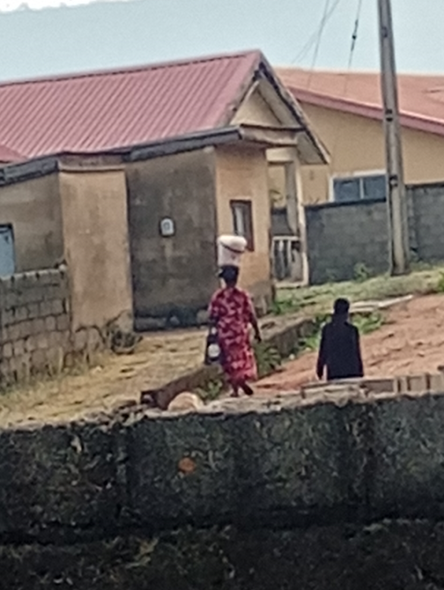 This image depicts a candid scene in Adankolo, Nigeria, under an overcast daytime sky. Two figures are visible in the mid-ground, against a backdrop of simple buildings.
The most prominent subject is a person, likely a woman, dressed in a red patterned garment, walking from left to right. She skillfully balances a white bucket or container on her head, a common practice for carrying goods in the region. She also appears to be carrying a bag in her left hand. Further to the right and slightly lower in the frame, a second individual, clad in dark clothing, is seen from behind, either standing or sitting by a low embankment.
The background features several single-story structures with light-colored, possibly plastered or painted, walls. Their roofs are distinctly reddish-brown and made of corrugated metal. One building has a small, gabled facade or porch structure. The ground beneath the figures and buildings is unpaved, dusty, and uneven, with patches of dry grass or weeds, characteristic of a semi-urban or rural environment. A dark, textured wall or embankment runs across the lower foreground of the image. The diffused light suggests it is daytime, without harsh shadows, possibly morning or late afternoon. No discernible text is visible anywhere in the image. The scene captures an everyday moment of activity within the community.