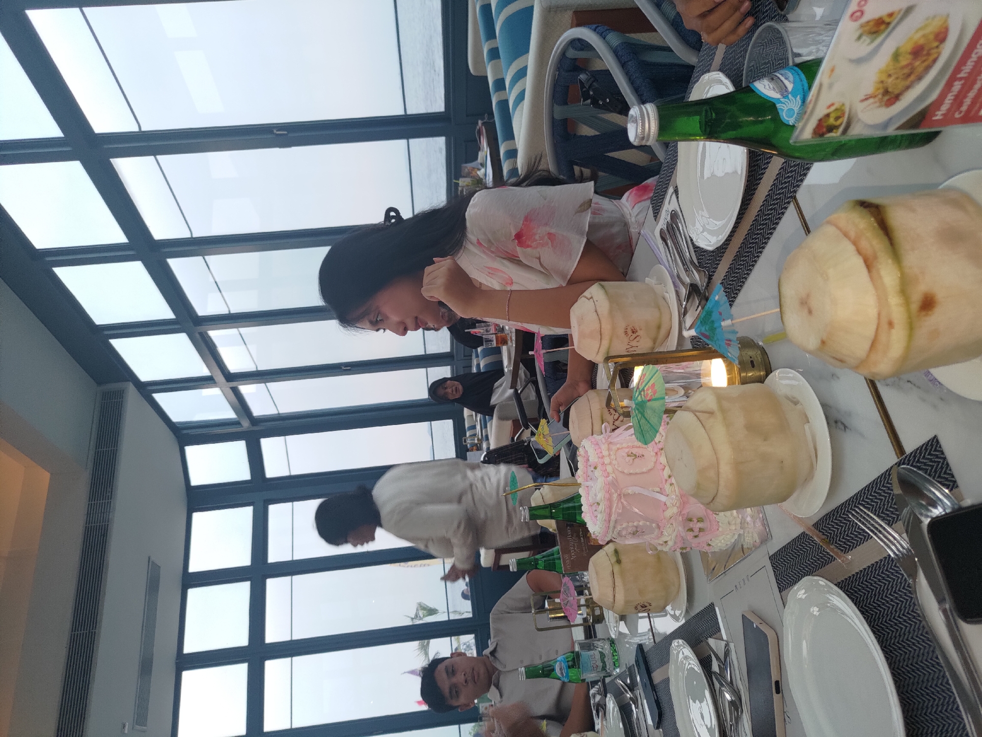 The image depicts a dining scene at a restaurant with large windows overlooking a body of water, likely the sea, suggesting a location in Kosambi Barat, Indonesia, known for its coastal proximity. The time of day appears to be late afternoon or early evening, with soft, diffused light filtering through the windows.
In the foreground, a table is laden with food and drinks, including several whole coconuts, a pink multi-tiered cake adorned with decorative icing and miniature umbrellas, and bottles of green liquid. Cutlery and plates are neatly set.
Several people are present. On the left, a young man in a light-colored collared shirt is seated, looking towards the camera with a neutral expression. To his right, a young woman with long dark hair, wearing a floral-patterned dress, is seated and looking to her left, with her hand resting on her chin, in a pensive pose. Behind the man, a woman in a white top is standing and gesturing with her hands, seemingly engaged in conversation. In the background, other diners are visible, including a person wearing a headscarf, seated at a table.
The restaurant interior features modern decor with blue and white striped seating and dark window frames. The overall atmosphere suggests a celebratory gathering or a relaxed meal by the waterfront. The presence of the cake and coconuts implies a special occasion, possibly a birthday or anniversary.