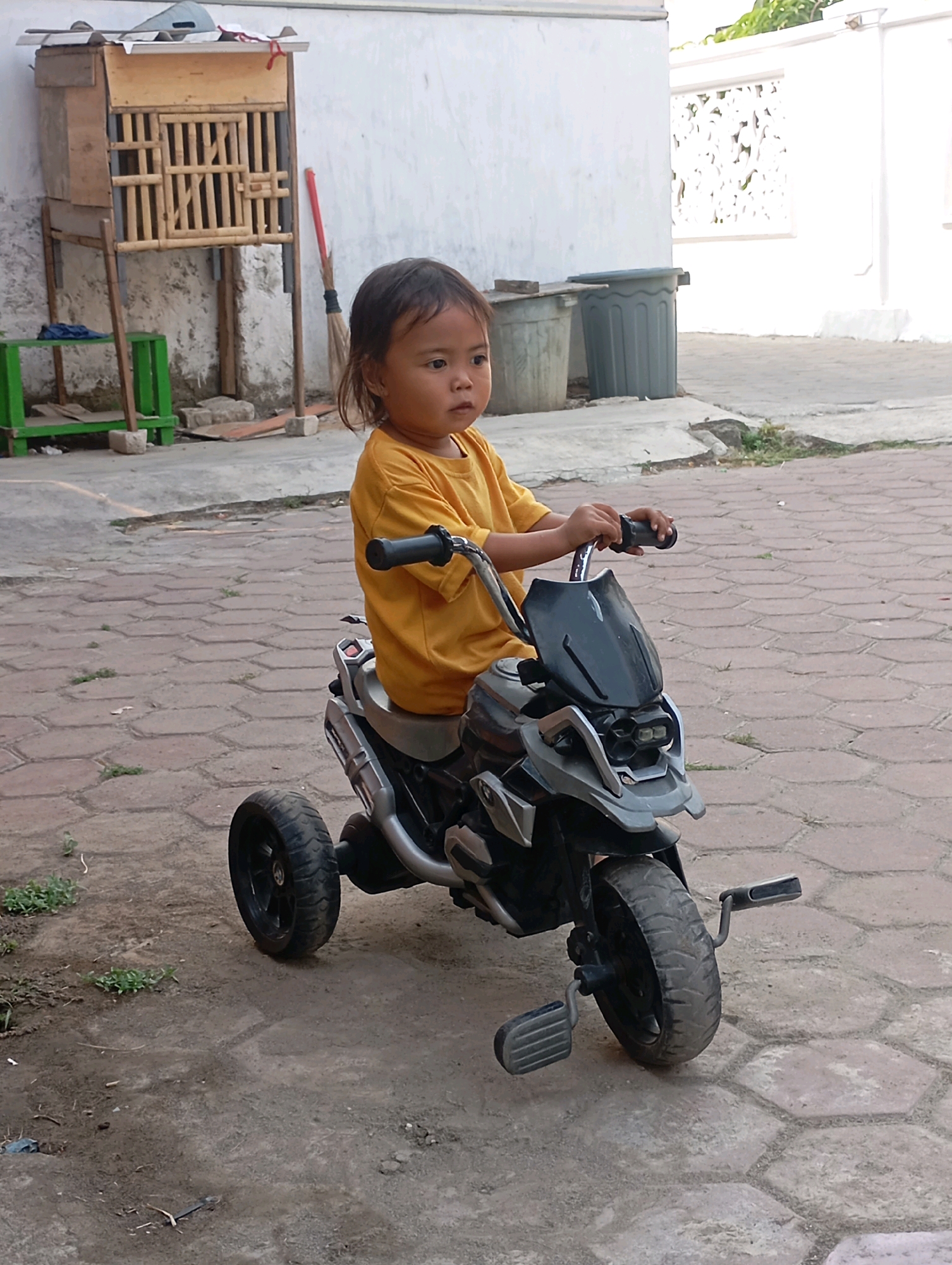 In Babelan, Indonesia, a young child with dark hair, wearing a yellow t-shirt, sits on a toy motorcycle. The motorcycle is dark gray and silver, with three wheels and a windshield. The child's hands are on the handlebars, suggesting they are operating the toy. The scene takes place on a paved area with a brick pattern, and a dirt area with sparse vegetation. In the background, a white wall is visible, along with a wooden structure that resembles a cage, a broom, and a gray trash bin. The overall environment appears to be an outdoor, residential area.