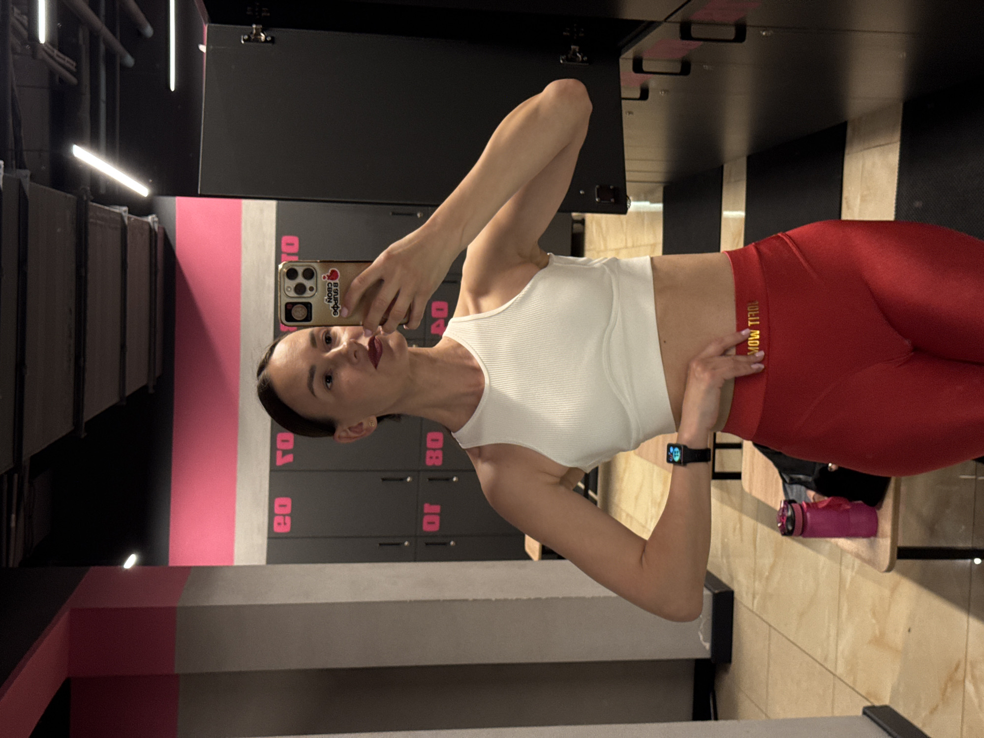 A woman takes a mirror selfie in what appears to be a gym locker room or changing area in Sochi, Russia. The image is presented sideways (rotated 90 degrees clockwise).
The woman, with dark hair pulled back, is wearing a white ribbed crop top and red high-waisted leggings. A black smartwatch with a colorful display is visible on her left wrist. She holds a gold-cased smartphone in her right hand, pointing it towards the mirror. The phone case features a red heart sticker and Russian text that reads "ВЛИЯЙ СВОИ" (VLIYAI SVOI), meaning "INFLUENCE YOUR". She has a slight pout and looks directly at the camera, conveying a confident demeanor.
The background reveals dark grey/black lockers, some marked with prominent pink numbers such as "08," "10," "20," and "80." Behind the lockers, a wall features a design of pink and dark grey stripes. The floor is covered with light-colored tiles, and the indoor space is illuminated by bright fluorescent strip lighting. On a light wooden bench or shelf to her right, a pink water bottle is placed. The waistband of her red leggings displays partially visible, mirrored gold lettering that appears to spell "GYM MODE." The overall environment indicates a fitness facility.