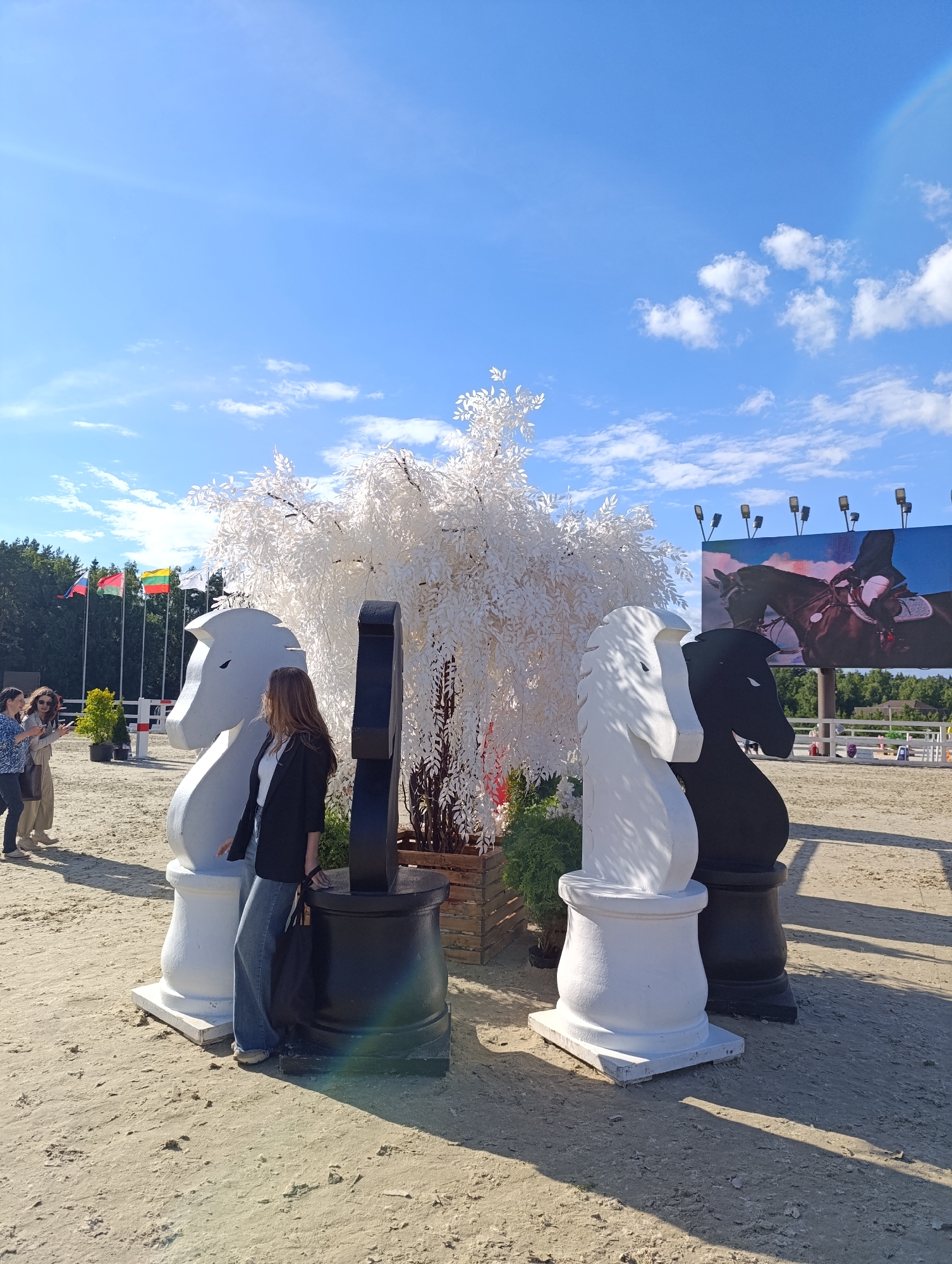 This is a full-shot outdoor photograph taken on a sunny day, featuring a sandy equestrian arena. In the foreground, a woman is standing between two oversized chess pieces, one white and one black, designed in the shape of horse heads. The white horse piece is to the left, and the black horse piece is to the right. Behind them is a large, decorative white tree with bare branches. In the background, several flags are visible on flagpoles, including the Russian flag. To the right, a large video screen displays an image of a horse and rider. Two other women are standing to the left of the main subject, one taking a picture with her phone. The ground is covered in sand, characteristic of an equestrian venue. The sky is blue with scattered white clouds.