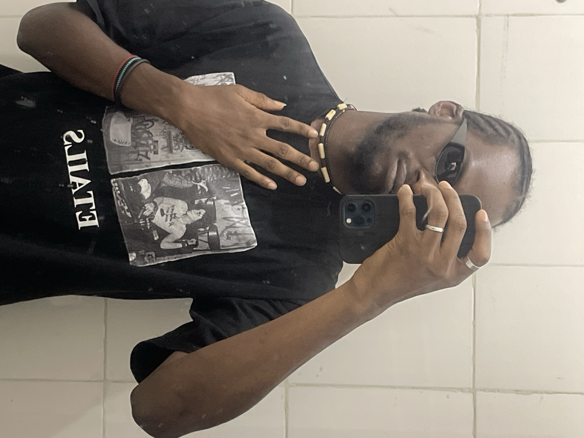 A male individual is captured in a mirror selfie in what is stated to be a location within Jos, Nigeria. He has braided hair and a short beard, wearing dark wraparound sunglasses that obscure his eyes. He is dressed in a black short-sleeved t-shirt featuring a graphic print of black and white images and mirrored text that appears to read "SJIATE" or "ETIALS" (reversed). A choker-style necklace with light-colored, rectangular beads strung on a dark cord is visible around his neck. His right hand holds a dark-cased smartphone with a visible camera module, positioned to take the photo; a silver-colored band ring is on his right index finger, and another on his right ring finger. His left hand is placed flat against his chest. A multi-strand beaded bracelet, featuring black, blue, and red beads, is on his left wrist. The background consists of a plain white tiled wall with visible grout lines.