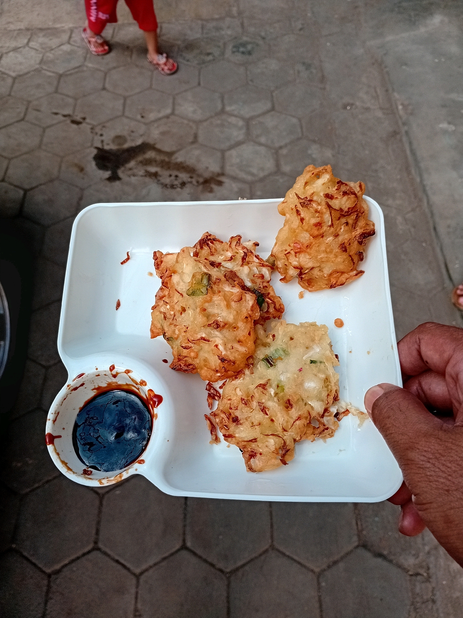 A close-up, overhead shot captures a white rectangular tray holding three pieces of fried fritters with a small cup of dark dipping sauce in the bottom left corner. The fritters, golden brown and irregular in shape, appear to be made from shredded vegetables, possibly carrots or potatoes, and green onions. The dipping sauce is thick and dark, possibly soy sauce or a similar condiment. A hand with dark skin is visible on the right side of the frame, holding the edge of the tray. The tray rests on a textured, hexagonal-patterned grey pavement. In the background, out of focus, a child's legs wearing red pants and patterned sandals are visible standing on the same pavement. A dark, wet stain is also present on the pavement in the background. The overall scene suggests an outdoor setting, possibly a street or courtyard in Gebang, Indonesia.