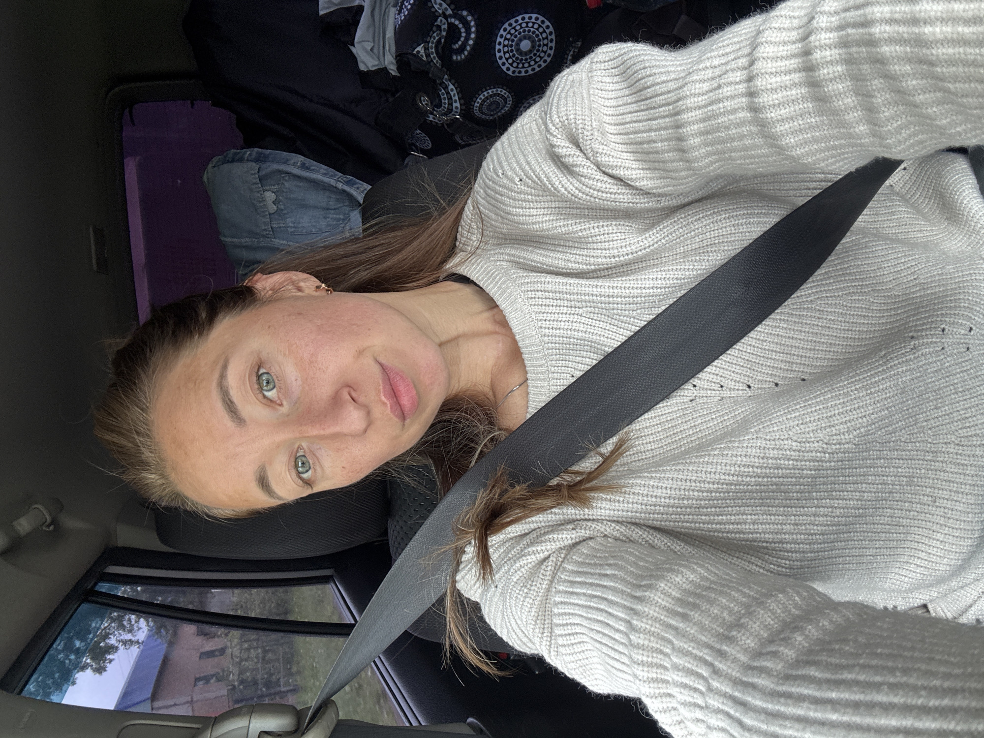 This image depicts a female individual seated inside a vehicle in Karagay, Russia. The subject has fair skin, light-colored eyes (appears blue-green), and light brown hair partially pulled back, with a small cluster of hair visibly caught in the seatbelt. She is wearing a light-colored (off-white or light grey) ribbed knit sweater, which exhibits minor distress or small holes, a small gold-colored hoop earring in her right ear, and a thin silver-colored necklace. Her expression is neutral with her lips slightly parted in a pout, and she is looking directly at the camera. A dark seatbelt crosses her chest and shoulder. Through the left side window of the vehicle, an exterior scene reveals a building with a reddish-brown facade and a prominent blue roof, followed by a smaller, darker building, and sparse vegetation under an overcast or twilight sky. The right rear window of the vehicle shows a purple tint with indistinct shapes in the background. Inside the vehicle, behind the subject, a folded denim jacket and a dark fabric bag with a white and grey circular pattern are visible on the car seats. The car's interior appears dimly lit.
