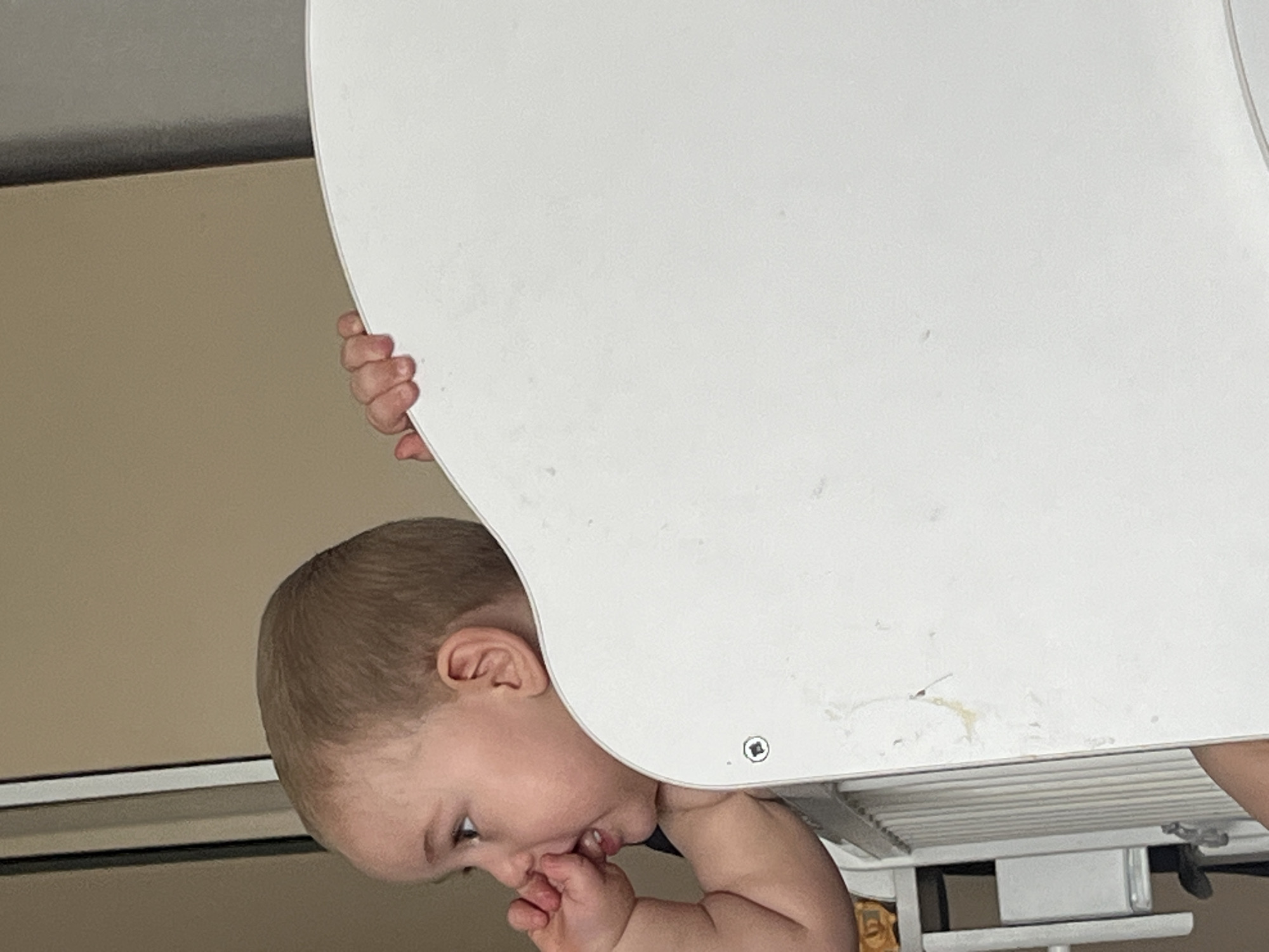 A toddler is peeking over the edge of a white object, possibly a crib or playpen. The toddler's left hand is on top of the white object, and its right hand is near its face, with a thumb in its mouth. The toddler is looking to the left with its mouth slightly open. The background is a plain, light brown wall. The image is taken indoors. The location in the city of Chelyabinsk, Russia, is not specified in the image.