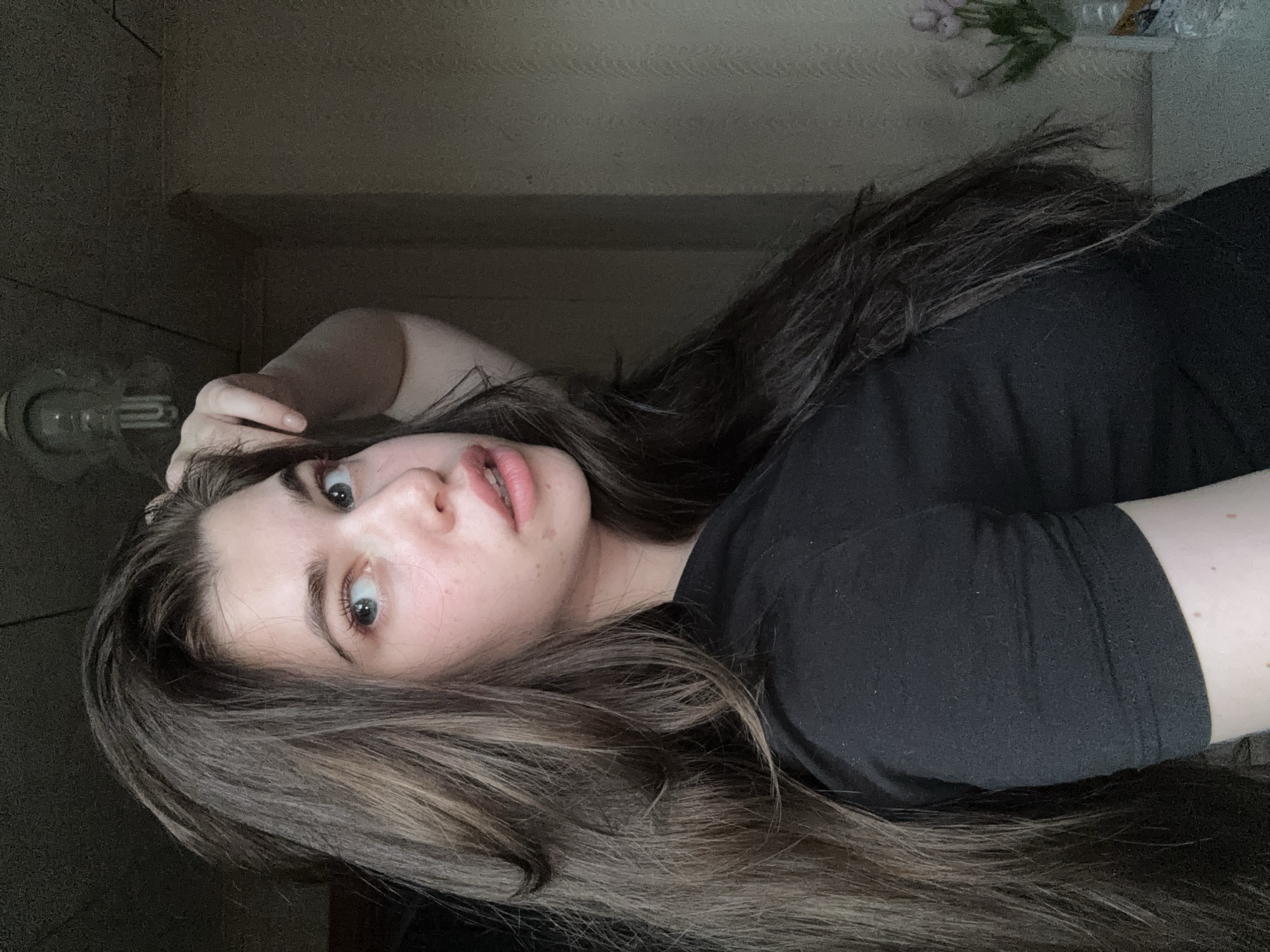 A woman with long brown hair is lying down and looking up at the camera. She is wearing a black t-shirt. There is a light fixture on the wall behind her and a vase with flowers on a shelf. The location is not identifiable as Мещёры, Russia from the provided image.