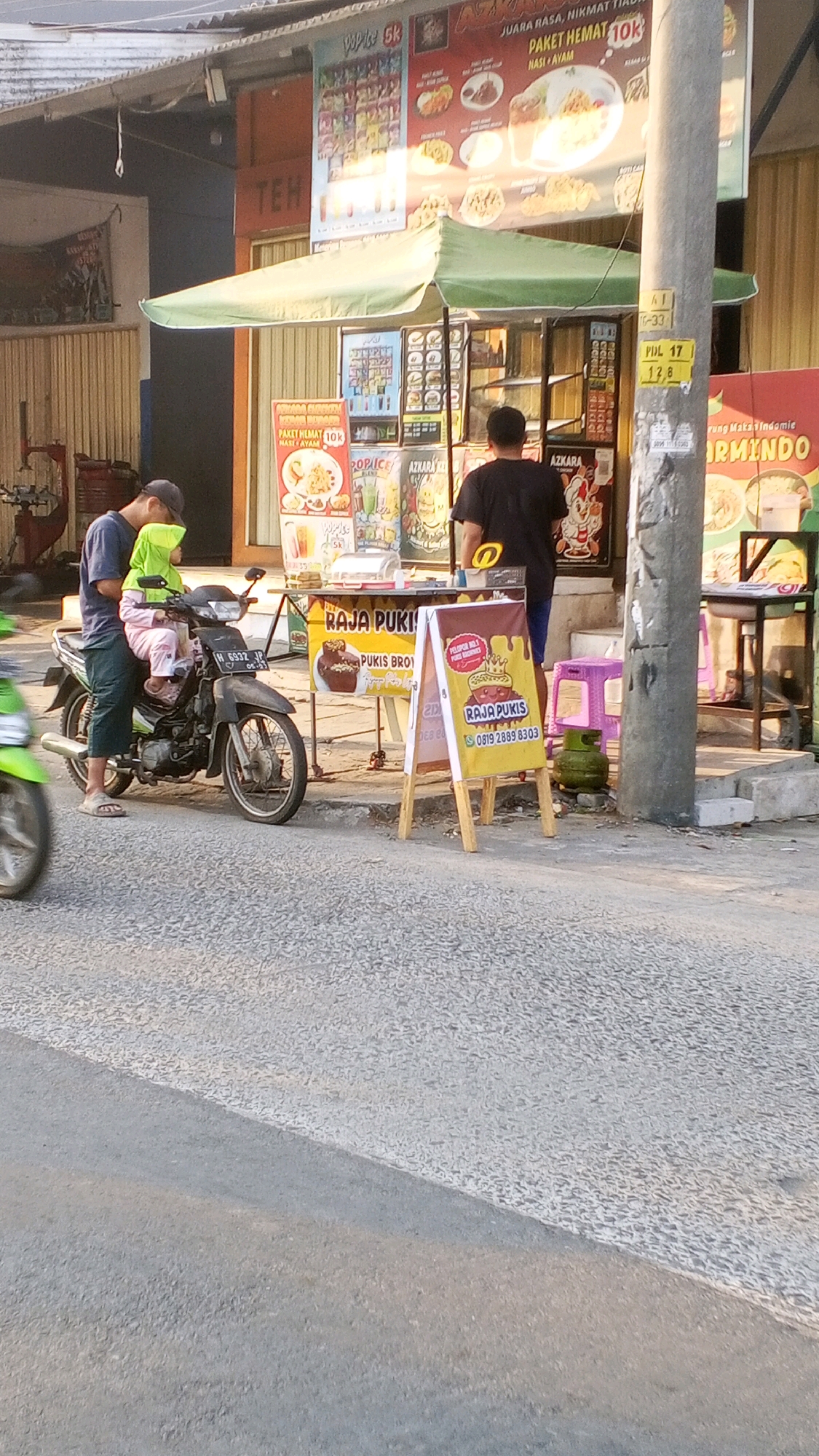 A man and a young child are seated on a black motorcycle parked on a roadside in Semarang, Indonesia. The man, wearing a dark blue t-shirt and a dark baseball cap, is on the left. The child, in a bright green hijab and light pink clothing, is on the right, positioned in front of the man. The motorcycle is facing right, with its front wheel turned slightly left. In the background, there is a food stall with a green awning and various signage displaying food items and prices. One sign features the words "RAJA PU-KIS" and "PUKIS BRO." A grey pole stands to the right of the stall. The ground is paved with asphalt, and there's a slight incline. The overall scene appears to be an outdoor urban setting during daylight.