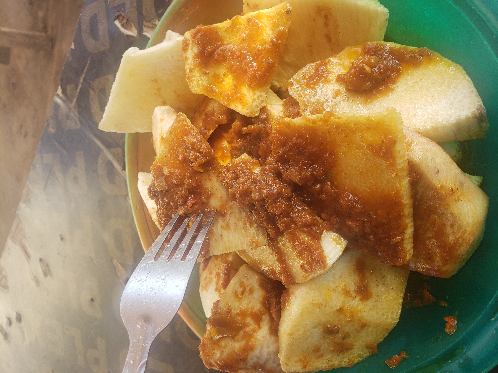 A fork is piercing slices of yam in a green bowl. The yam is topped with a brown sauce. The scene is set on a wooden table. The image is taken in Gumel, Nigeria.