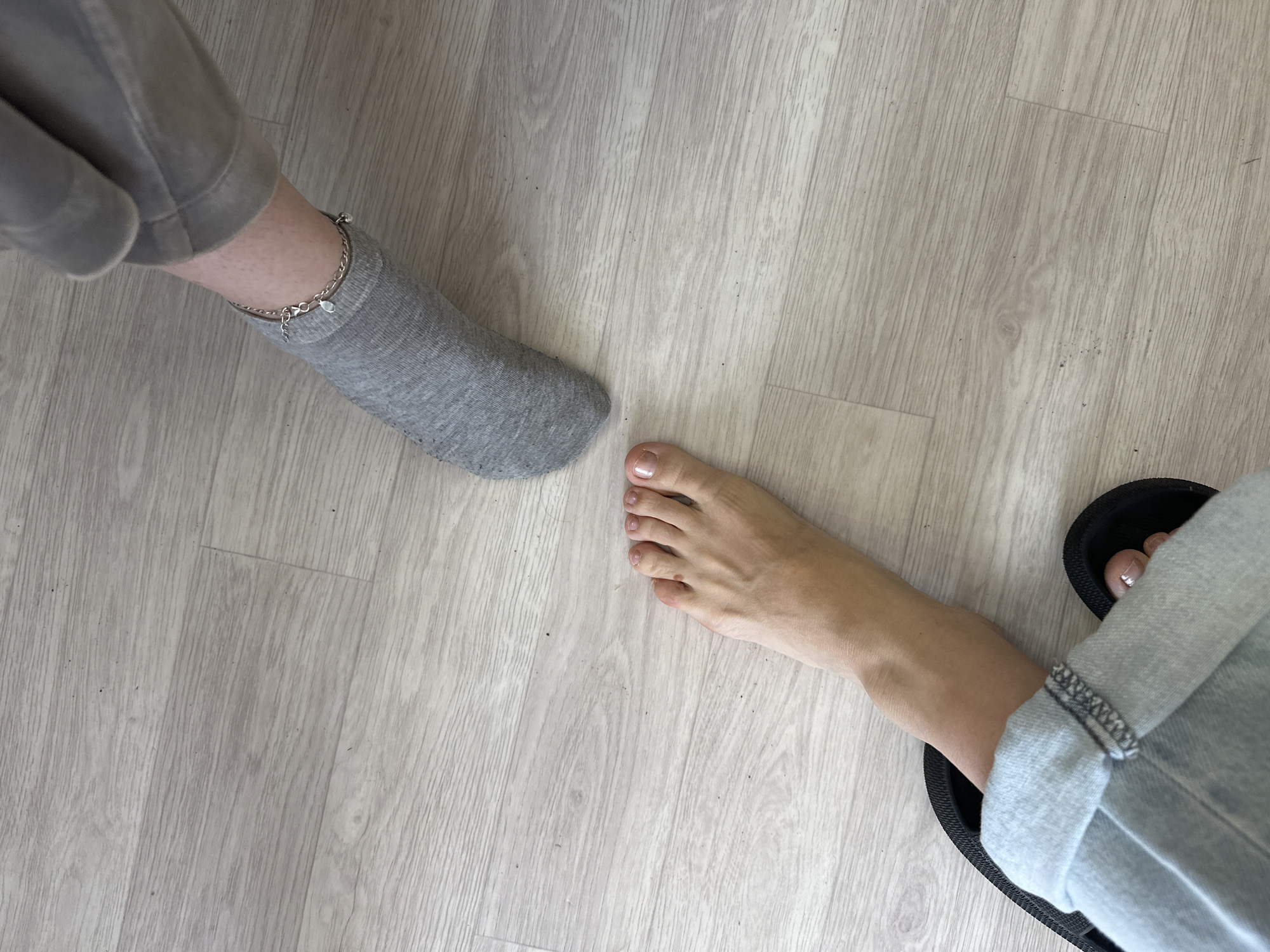 Two bare feet are visible on a light-colored wooden floor. One foot, on the left, is positioned at the bottom of the frame, wearing a light blue cuffed jean and a black sandal. The toes are visible and appear to have clear nail polish. The other foot, on the right, is wearing a grey sock and a silver anklet with a small charm. This foot is positioned higher in the frame, with the leg covered by grey sweatpants. The floor has a visible wood grain pattern with a few small dark specks scattered across it.