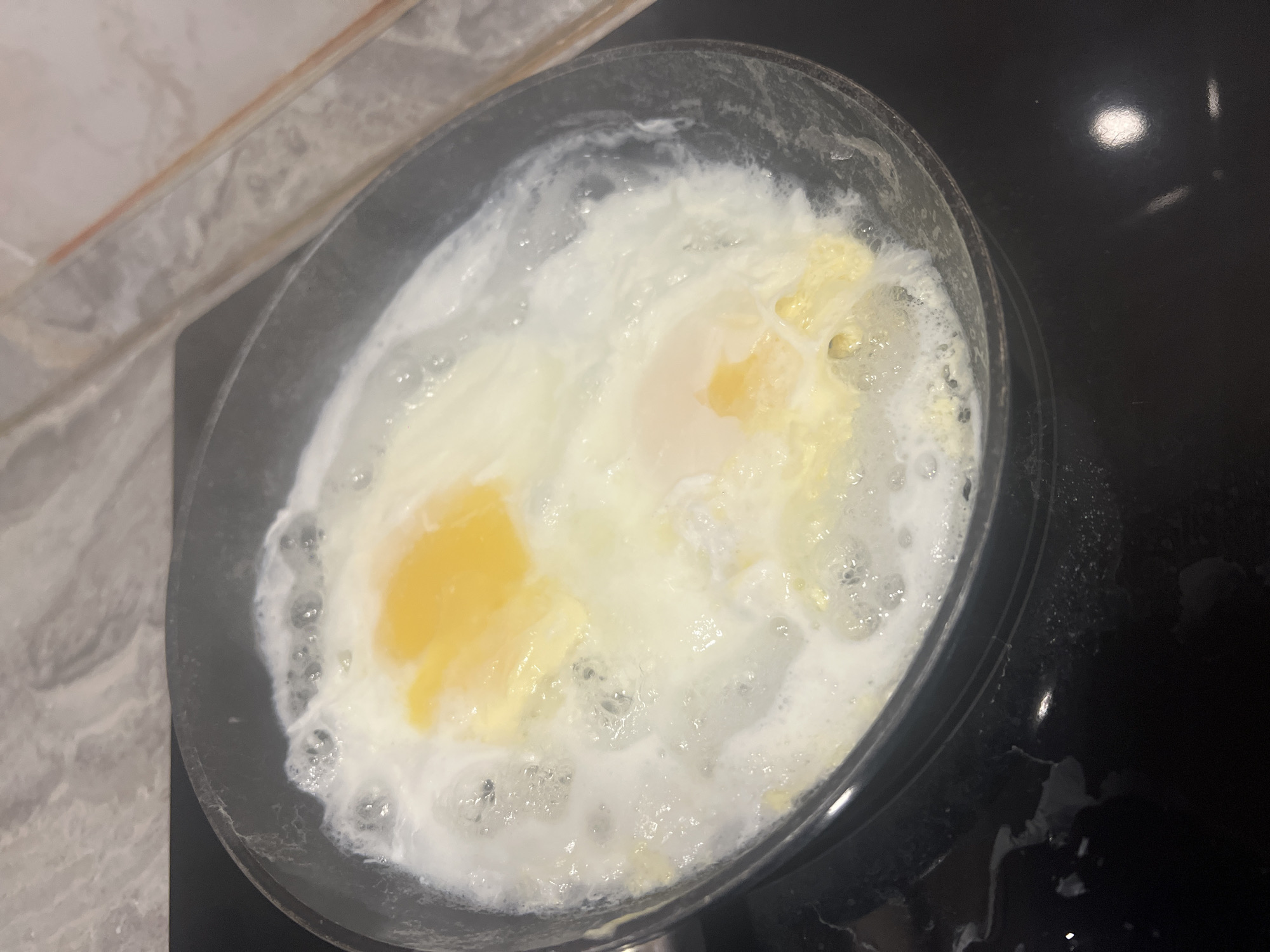 A close-up shot shows two eggs frying in a black pan, placed on a black stovetop. The egg whites are mostly cooked, with visible bubbles and some solidified areas. The egg yolks are still runny and a vibrant yellow. The edge of the stovetop and the pan's side are visible on the right. A countertop with a marble-like pattern is seen on the left.