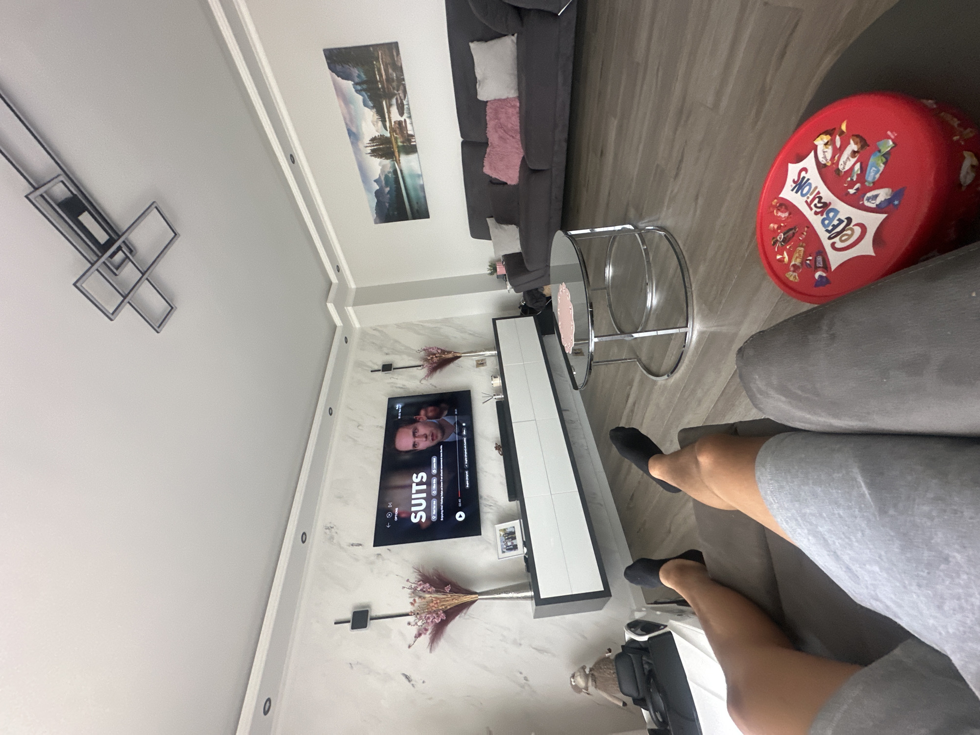 The image depicts an indoor scene, specifically a modern living room located in Marinka, Bulgaria. The perspective is low, showing the legs of an individual in grey shorts and black socks, resting on a grey fabric sofa in the foreground. To the right of the individual's legs, on a wood-effect laminate floor, is a red oval-shaped "Celebrations" candy container.
In the midground, a round glass coffee table with a polished chrome base is visible. Beyond this, a dark grey three-seater sofa, adorned with one pink, two light grey, and one dark grey cushion, is positioned against a plain white wall on the right side of the room. Above this sofa, a wide panoramic print featuring a mountain and lake landscape is mounted.
The left side of the room features a prominent accent wall with a white and grey marble-effect finish. A large flat-screen television is mounted centrally on this wall, displaying a scene from "SUITS" with a male actor's face visible, along with user interface elements like play/pause controls and language options. Beneath the television, a floating console unit, comprising dark grey sides and white drawer fronts, is mounted. On the console, from left to right, are a tall silver vase holding dried pink and beige floral arrangements, a small framed photograph, and a black soundbar. Another identical floral arrangement in a silver vase is positioned on the right side of the console. Two slender, black wall-mounted fixtures are vertically positioned on the accent wall, flanking the television. In the far left corner, a light brown plush sheep toy is visible, resting on a white and black object, possibly a child's seat or playpen.
The ceiling is white and features recessed lighting fixtures along the perimeter. A contemporary, geometric chandelier with a black frame and integrated lighting is centrally mounted on the ceiling, further towards the perspective of the shot. White crown molding delineates the transition between the walls and the ceiling throughout the room. The overall environment is well-lit and appears tidy.