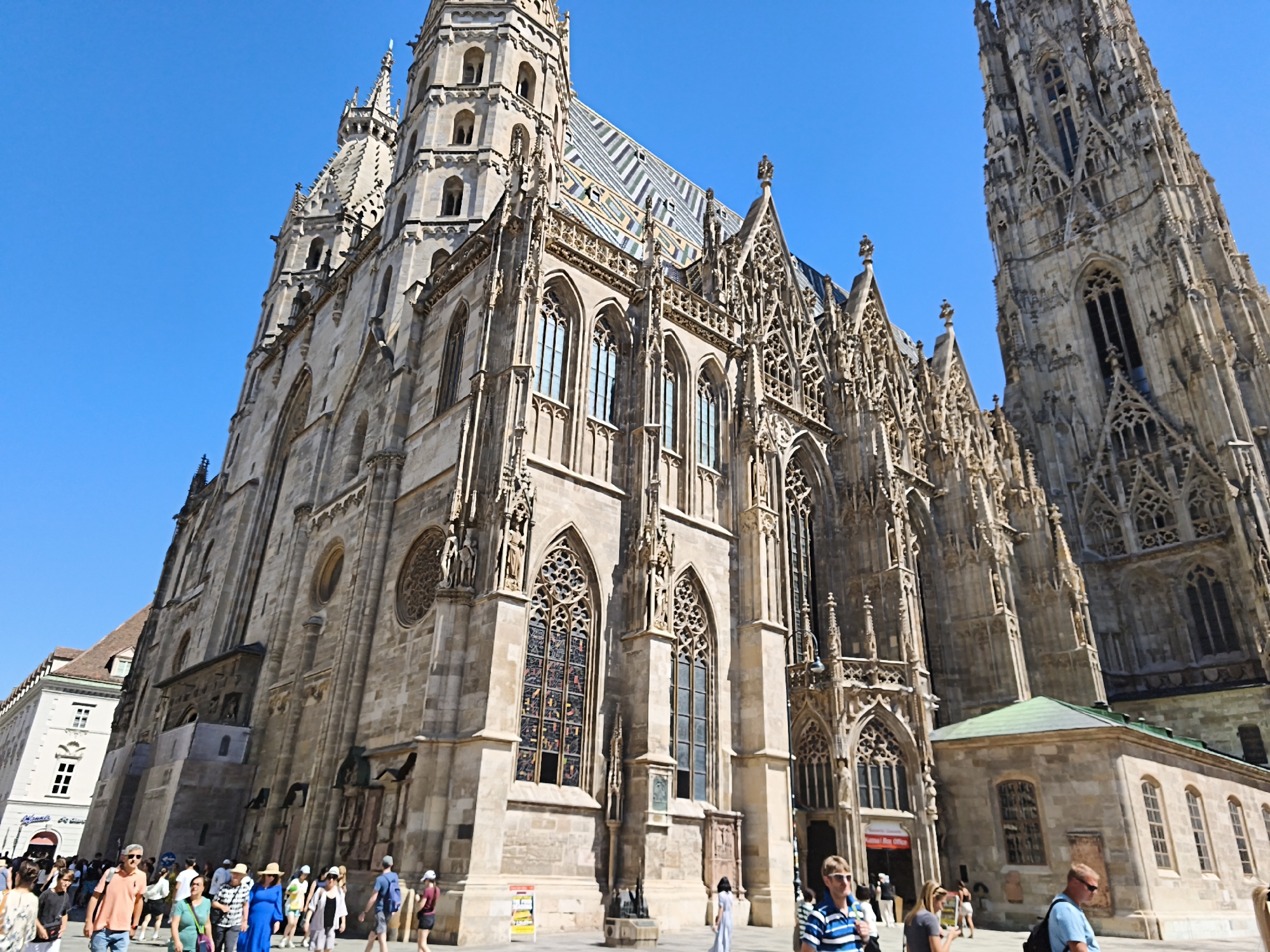 In Vienna, Austria, a low-angle shot captures the imposing St. Stephen's Cathedral under a clear blue sky. The Gothic architecture is detailed with ornate carvings, flying buttresses, and intricate window tracery. The cathedral's steeply pitched roof is covered in a distinctive patterned tile mosaic. In the foreground, a crowd of tourists mills about on a paved plaza. People are dressed in casual summer attire, some are walking, others are gathered in small groups. Among them is a man in a blue and white striped polo shirt, a woman in a blue dress, and individuals wearing t-shirts and shorts. A small, green-roofed building abuts the cathedral on the right. The overall scene depicts a popular tourist attraction on a sunny day.
