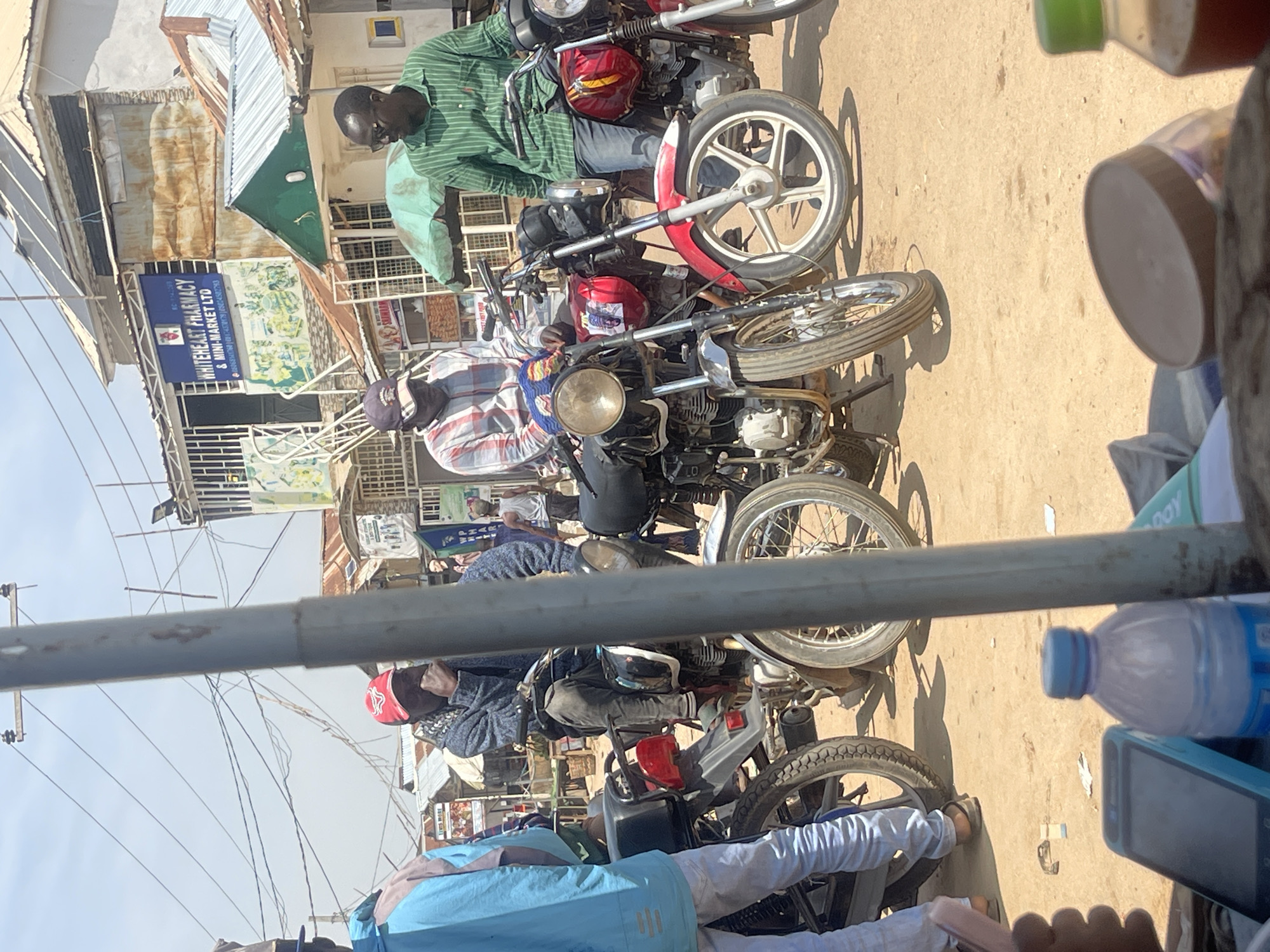 The image shows a street scene in Abuja, Nigeria, with several people and motorcycles. The individuals are dressed in casual clothing, suggesting a warm day. Two men are seated on motorcycles, one in a green and white striped shirt, and another in a checkered shirt. Another person is standing behind them, wearing a blue jacket. The motorcycles appear to be common models used for transportation in the region, with one red and white bike prominent in the foreground.
The setting is an outdoor market or busy street, indicated by the buildings in the background. One building has a sign that reads "WHITEHEART PHARMACY & MINI-MARKET LTD". Another sign displays colorful images. Utility poles and wires are visible, suggesting the area is urban. The ground is dusty and appears to be unpaved in some areas. The sunlight casts shadows, indicating it is daytime.
In the foreground, a person in a light blue shirt and white pants is partially visible, along with another person wearing white pants. A water bottle and a smartphone are also in the foreground. The overall impression is of a bustling everyday scene in a Nigerian city.