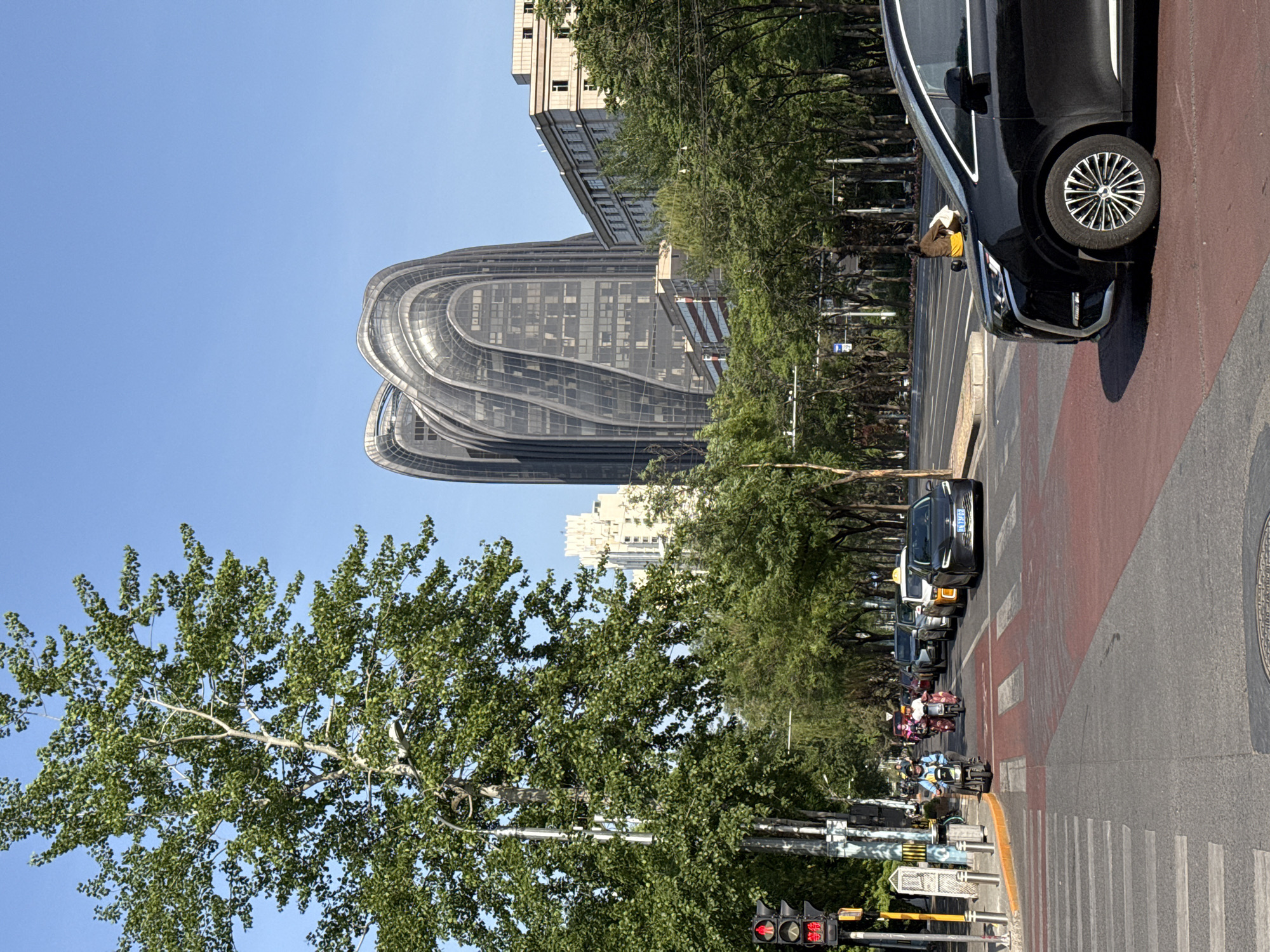 The image captures a street scene in Chaoyang District, Beijing, China. A distinctive architectural structure, possibly a modern office building with a flowing, curved design, dominates the background. The sky is clear and blue. The street in the foreground has crosswalks. A row of parked cars lines the right side of the street, one black car being particularly prominent. A mix of motorized scooters and bicycles populate the road, suggesting urban traffic. A tall tree with green leaves frames the left side of the view. A traffic light is visible in the lower left corner. The scene conveys an urban environment with modern architecture and busy street activity.