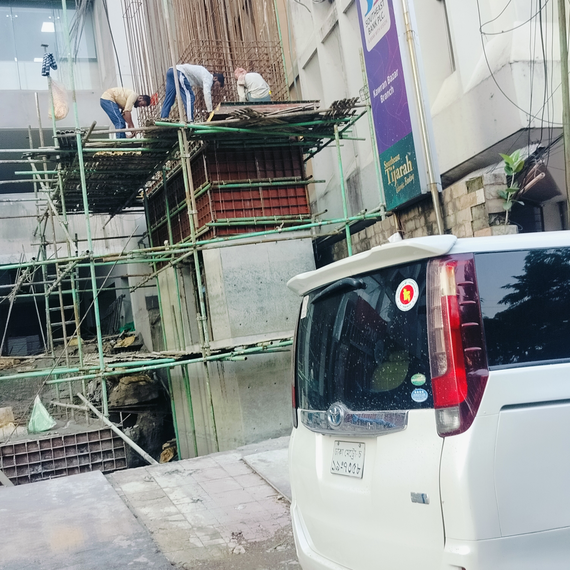 The image depicts a construction site in Dhaka, Bangladesh, featuring three workers on scaffolding. The site appears to be adjacent to the "Southeast Bank PLC., Kawran Bazar Branch". A white minivan with the license plate "ঢাকা মেট্রো-চ ১৯-৭৩৫৮" is parked in the foreground, partially obscuring the view. The vehicle has a sticker of the flag of Bangladesh on its back. The scaffolding is constructed of green pipes and planks, with metal reinforcement bars visible within the construction itself.
