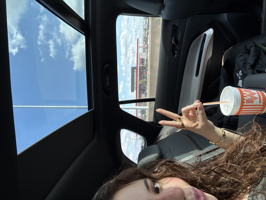 Inside a car with a panoramic sunroof revealing a blue sky with clouds, a person holds up two fingers in a peace sign. They also hold a Whataburger drink with an orange straw. The car interior is black and gray. Outside the window, a large building with red accents is visible. The person has brown curly hair and wears gold jewelry, including rings and a bracelet.