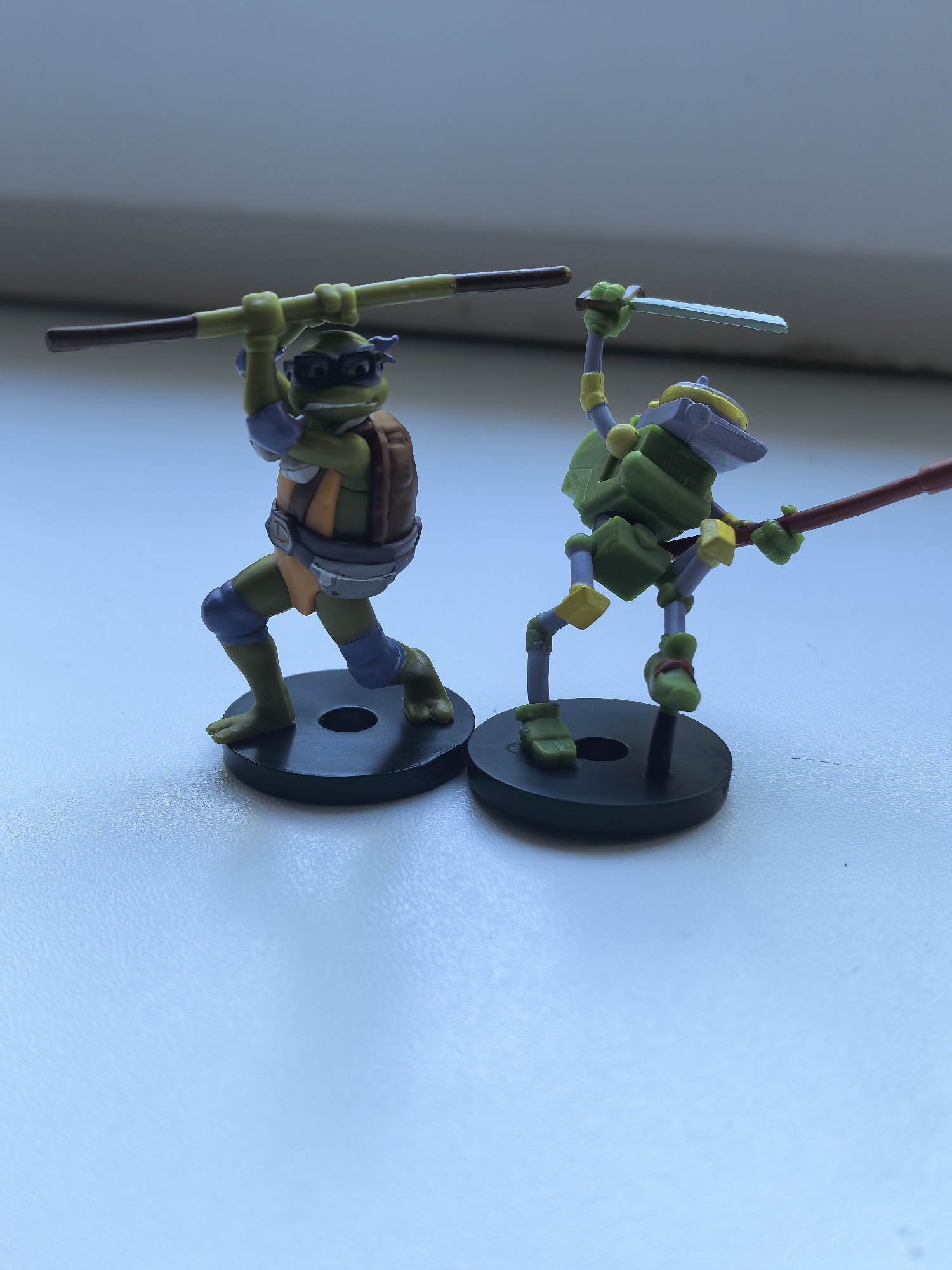 Two Teenage Mutant Ninja Turtles action figures stand on a white surface. The figure on the left is Donatello, wielding his bo staff. The figure on the right is a robotic turtle, wielding a sword in one hand and a bo staff in the other. Both figures are standing on black circular bases. The background is blurry but appears to be a windowsill with natural light. The image was not taken in Sterlitamak, Russia.