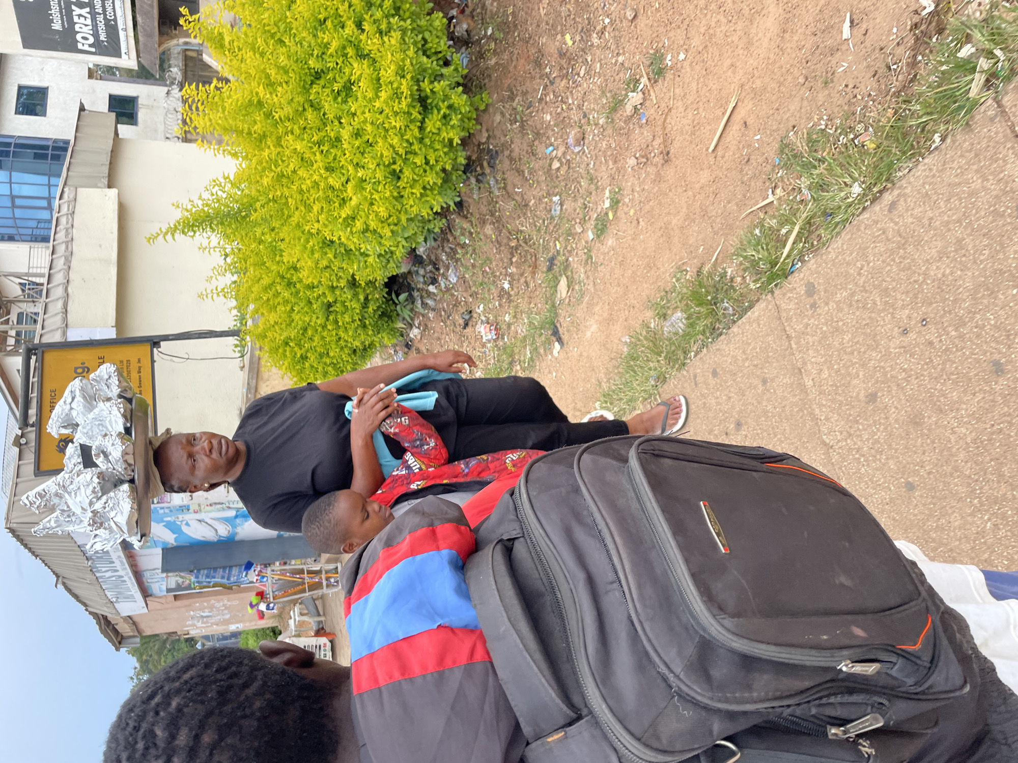 An outdoor, eye-level, full shot captures a woman in Jos, Nigeria, walking on a sidewalk. She is carrying a tray of foil-wrapped items on her head and a child is strapped to her back, wearing a red jacket with a cartoon print. The woman wears a black t-shirt and black trousers, with white flip-flops on her feet. She holds a light blue cloth in her right hand. In the foreground, the back of a person wearing a black backpack is visible. The background includes a building with signage, lush green bushes, and a dirt path with scattered debris. The sky is overcast.