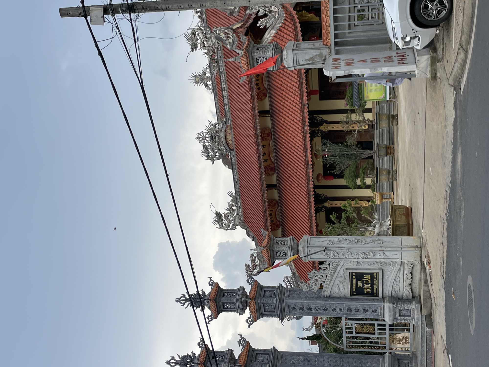 The image shows the entrance to the Truc Lam Zen Monastery in Da Lat, Vietnam, a Buddhist temple set against a cloudy sky. The temple features traditional Vietnamese architecture with red tiled roofs, ornate dragon sculptures along the eaves, and intricately carved decorative elements. The entrance is flanked by two large stone pillars with detailed carvings and decorative finials. A metal gate is visible behind the pillars, and a sign on the pillar reads "CHÙA TRÚC LÂM".
In front of the temple, there are several potted bonsai trees, adding to the serene atmosphere. A white car is parked to the right of the frame. A person in light-colored clothing, possibly a monk, is seen walking inside the temple grounds. The street in front of the temple is paved, with a manhole cover visible in the foreground. The overall scene captures the architectural beauty and spiritual ambiance of the monastery.