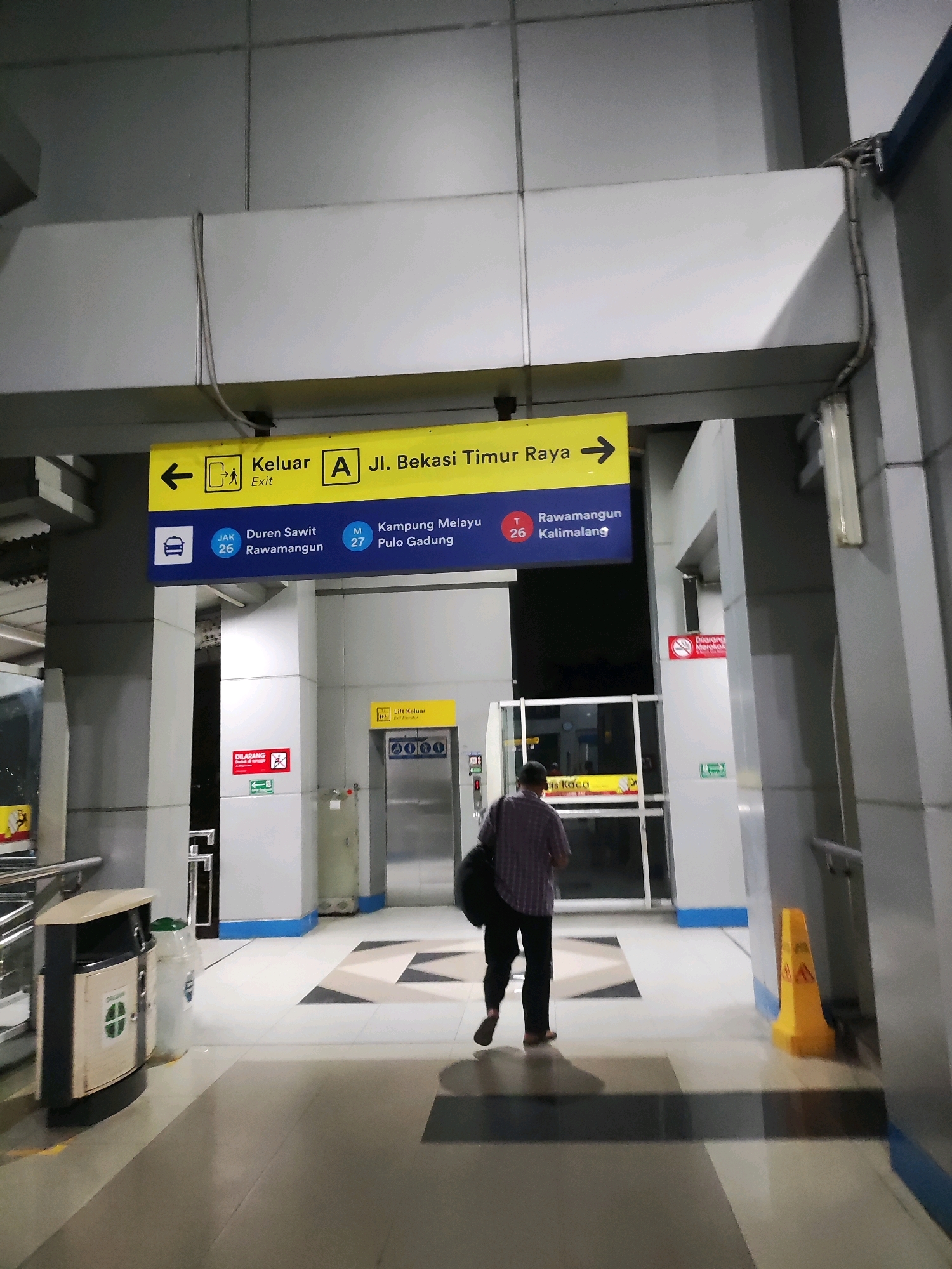 The image captures an indoor view of a station's platform in East Jakarta, Indonesia, likely an LRT or train station. A person, wearing a checkered shirt and dark pants, walks away from the camera towards the right side of the frame. Above them, a yellow directional sign indicates "Keluar Exit" with an arrow pointing left, and "A Jl. Bekasi Timur Raya" with an arrow pointing right, suggesting this is a main artery. Below this, a blue sign lists various destinations with alphanumeric codes, including "JAK 26 Duren Sawit Rawamangun" and "M 27 Kampung Melayu Pulo Gadung," and "T 26 Rawamangun Kalimalang." To the left, a trash can and a lighter-colored bin are visible. To the right, a yellow caution cone with warning symbols is positioned on the floor. Further down the hall, an elevator with the text "Lift Keluar Exit" is visible. The environment is constructed with metallic, grey, and white panels, with a patterned floor. The lighting appears to be artificial, casting a slight yellow hue. Red and green signs indicating prohibitions and emergency exits are also present on the walls.