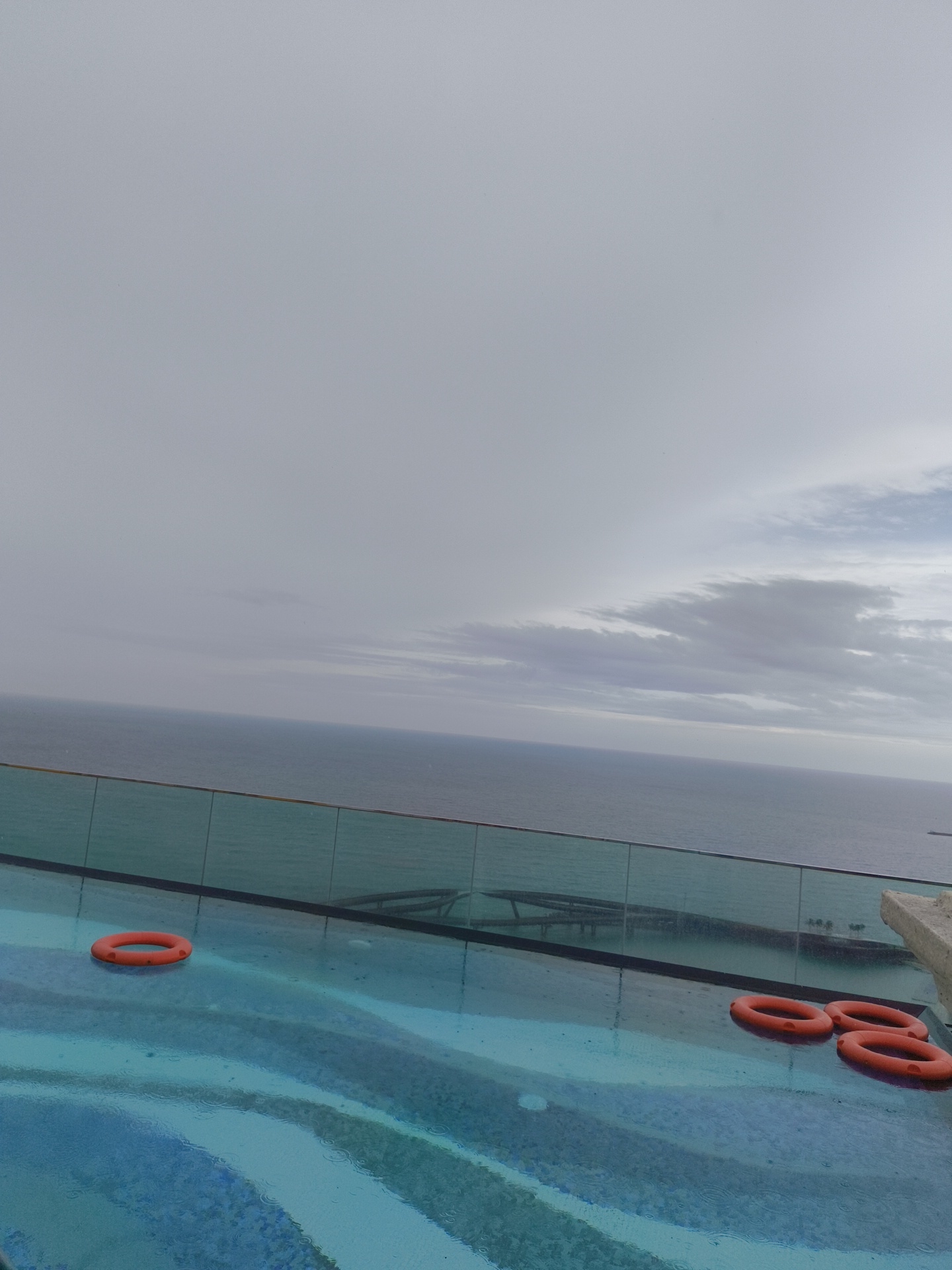 The image depicts an outdoor infinity pool with a glass barrier, overlooking a calm, gray ocean under an overcast sky. Four orange life preservers are floating in the pool. One is on the left side, and three are clustered on the right. The pool's surface shows ripples from rain. The location is identified as the city of Phú Quốc, Vietnam. The scene is devoid of people and any visible activity.