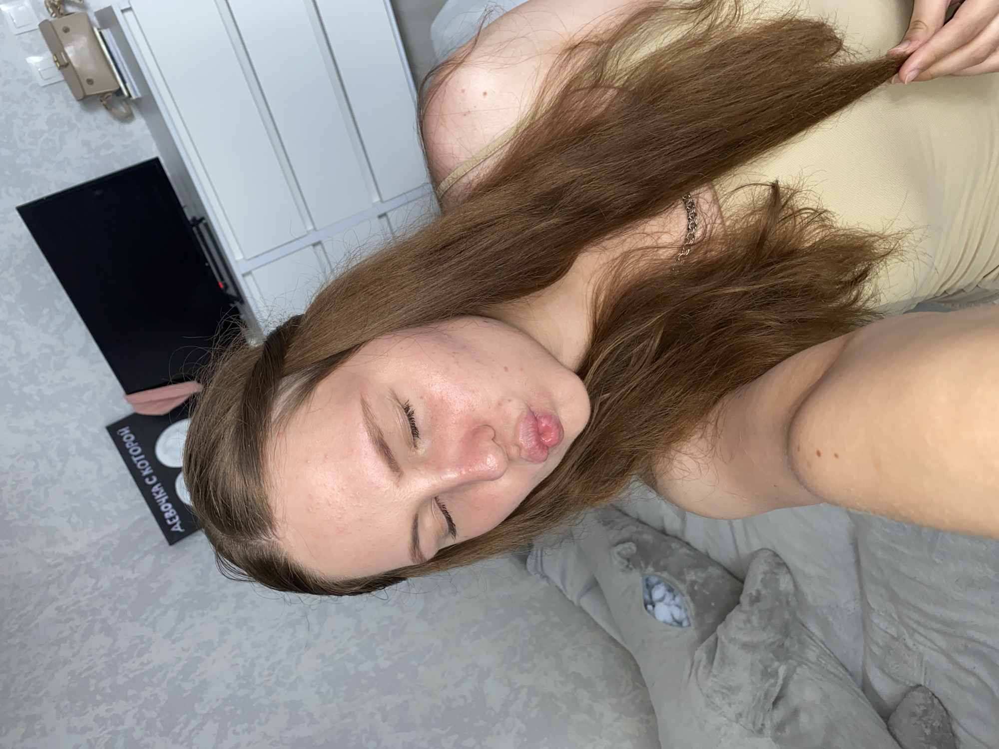 A young woman with long, brown hair is taking a selfie in a bedroom. She has her eyes closed and her lips pursed as if blowing a kiss. She is wearing a light-colored tank top. In the background, there is a large grey stuffed animal, a white dresser with a black television on top, and a framed picture on the wall with Russian text. The room appears to be wallpapered with a grey textured pattern. There are no clear indicators of the specific location within Izhevsk, Russia.