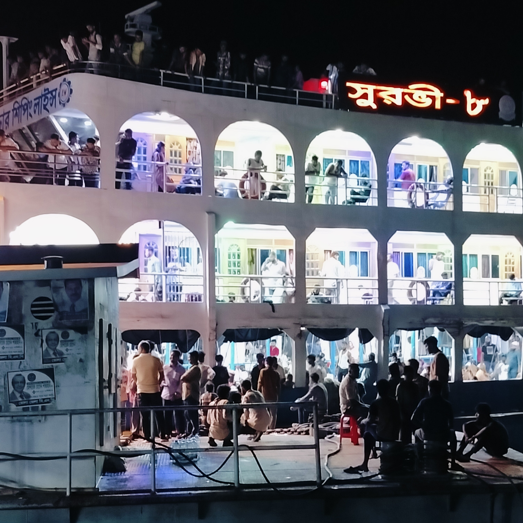 The image depicts a large, multi-deck passenger ferry, likely docked or stationary at night, bustling with numerous people. The dark night sky highlights the brightly illuminated interior and exterior of the vessel, which glows with a mix of warm and cool artificial lights from its arched windows and open areas.
On the uppermost deck, many people stand along the railing, observing their surroundings. A radar or antenna structure is visible at the very top. Below this, the side of the boat clearly displays "ডবল স্টার শিপিং লাইন" (Double Star Shipping Line) in Bengali script.
The middle decks feature multiple cabins or lounges, each with arched openings and brightly lit interiors. People are seen inside these areas, some standing by the railings or windows, while others appear to be seated or moving within the cabins. A lifebuoy is visible within one of these sections.
The lowest visible deck, closest to the viewer, is densely populated with men. Some are standing, while others are seated on improvised arrangements like coiled ropes, hoses, or a red plastic stool, suggesting a period of waiting or informal social gathering. Electrical wires or cables are present on this deck. On a structure to the left of this deck, several posters with faces of men and Bengali text are affixed. These appear to be campaign or promotional posters, with phrases like "দেশের জনগন" (people of the country) and dates potentially visible.
Dominating the top right of the ferry, large, illuminated red Bengali characters spell out "সুরভী - ৮" (Surabhi - 8), indicating the vessel's name and number.
The overall scene conveys a sense of busy transit or departure/arrival activity typical of passenger ferries in regions like Bhola, Bangladesh, where river and coastal transport are vital. People appear calm, engaged in waiting, conversing, or observing the night.