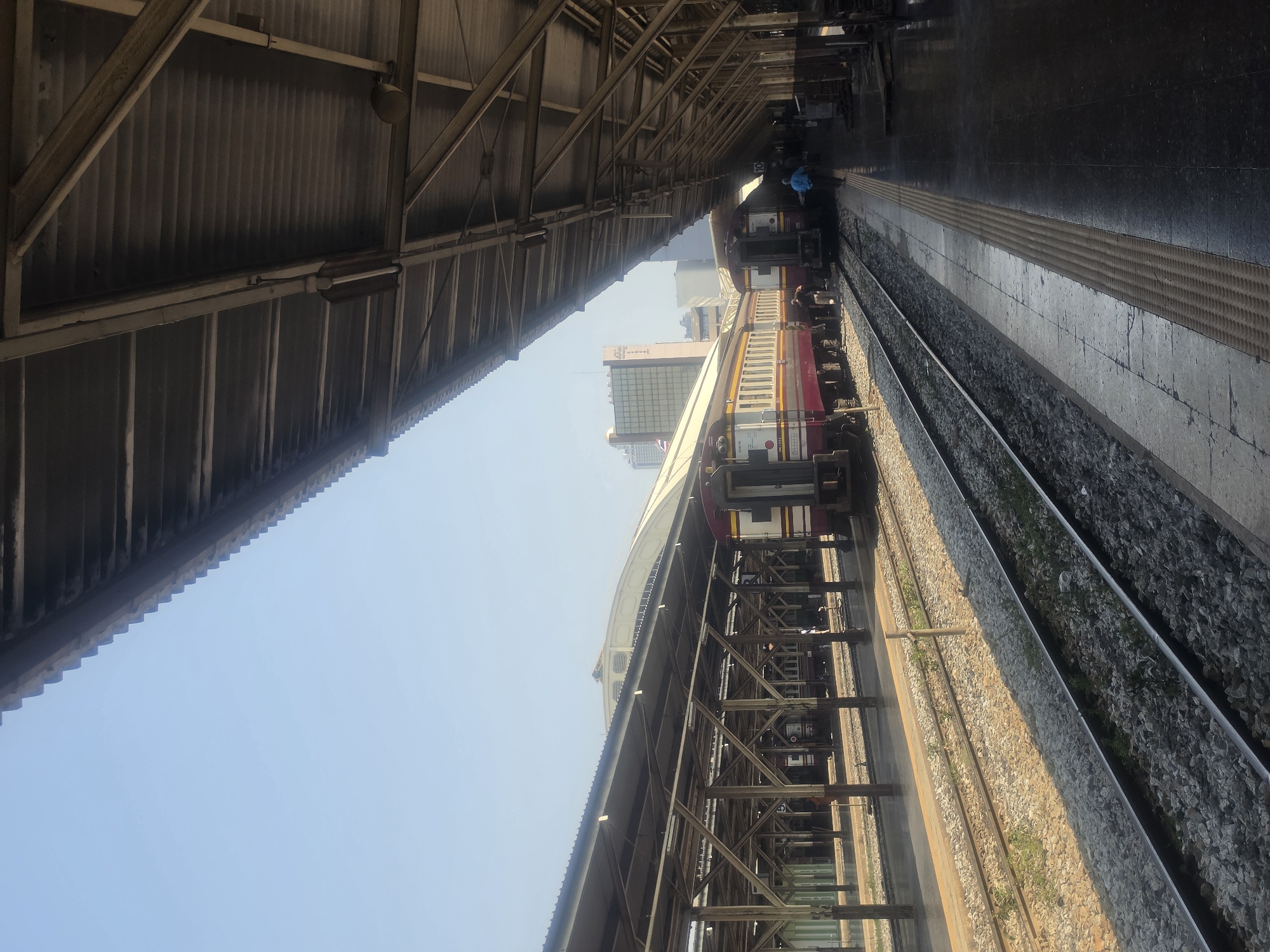 This image captures a vibrant daytime scene at a train station in Bangkok, Thailand, viewed from under a platform roof. The perspective is tilted, with the station canopy forming a prominent diagonal element.
On the left, the underside of a corrugated metal roof is visible, supported by a sturdy framework of metal beams and trusses. Attached to these beams are a round light fixture and a fluorescent tube light.
To the right, a train is stopped at a platform. Its carriages feature a distinct livery of red, cream, and yellow/orange stripes, characteristic of State Railway of Thailand (SRT) trains. Several indistinct figures can be seen on the platform beside the train, one notably wearing a blue shirt. Beyond the platform, multiple parallel train tracks are laid on gravel ballast, with small areas of greenery growing between them.
The background reveals a clear, bright blue sky, and in the distance, a modern city skyline with towering buildings and glass facades is visible, underscoring the urban setting. The bright illumination suggests it is mid-morning or afternoon.
Visible text on the train carriages includes partial alphanumeric codes and numbers, such as "SRT" and what appears to be car identification numbers like "1110" or "1141." The overall setting, train design, and city architecture strongly point to the specified location of Bangkok, Thailand.