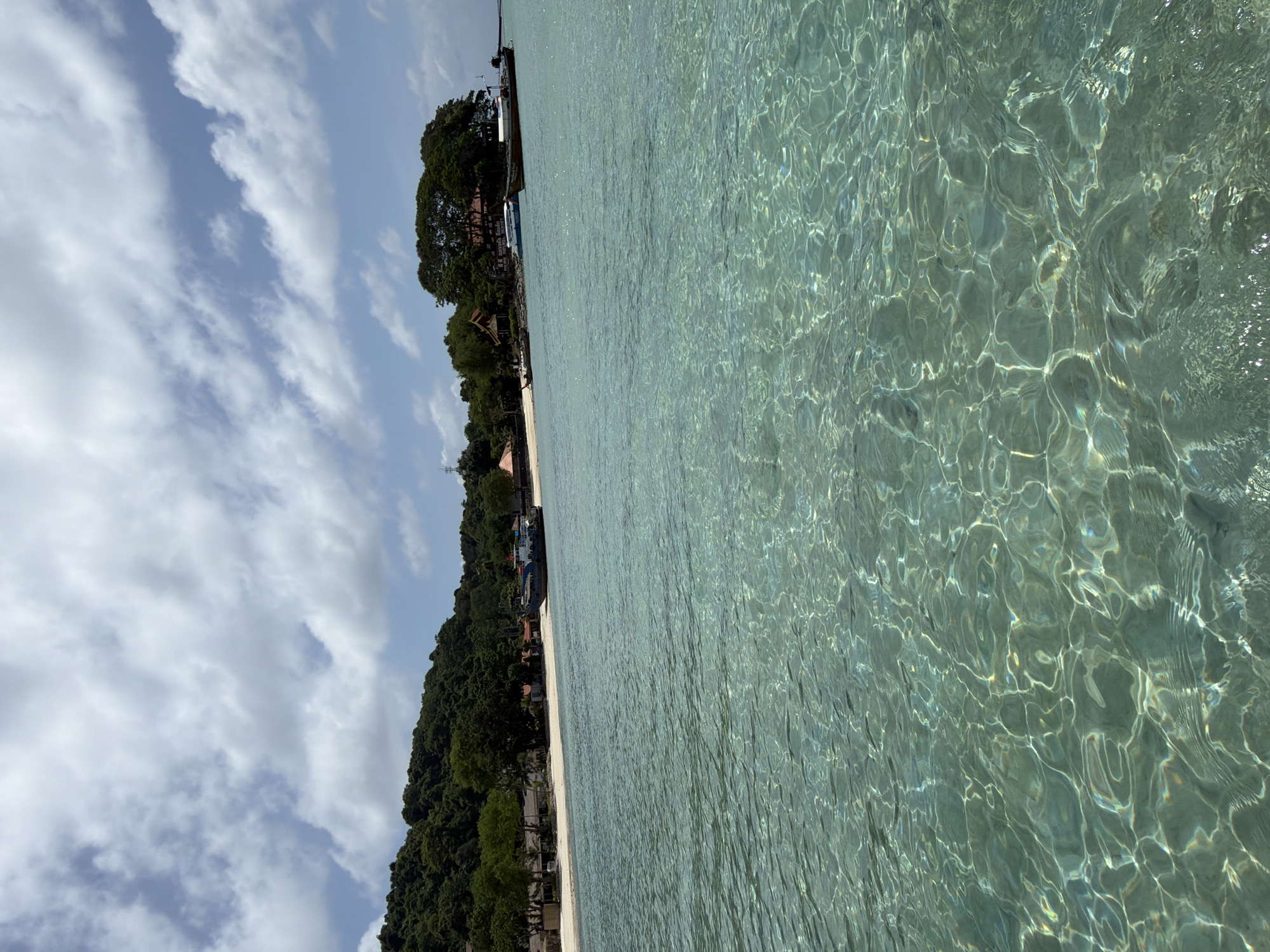 The image depicts a tranquil scene of a tropical beach in Ao Nang, Thailand. The foreground is dominated by crystal-clear turquoise water, with sunlight creating shimmering patterns on the seabed. In the mid-ground, a white sandy beach stretches along the coastline, backed by lush green vegetation and a small cluster of buildings. Several boats are anchored near the shore, including a longtail boat and a larger vessel. The background features a verdant, tree-covered hillside rising towards a bright blue sky filled with scattered white clouds. The overall atmosphere is one of serene natural beauty and coastal relaxation.