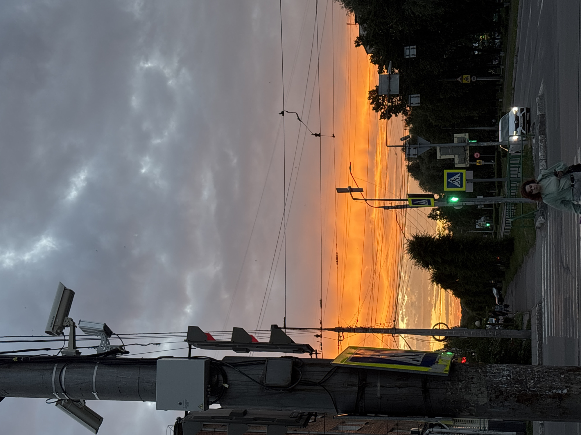 The image depicts a street scene in Novokuznetsk, Russia, at dusk. A concrete utility pole, equipped with surveillance cameras and traffic lights, stands prominently in the foreground on the left. The sky is dramatic, with a vibrant orange and yellow sunset illuminating the clouds.
Several traffic signals are visible, including one with a green light indicating it is safe to proceed. A pedestrian crossing is marked on the asphalt road. A woman in a light green top is walking across the pedestrian crossing, holding a smartphone. Further down the street, a white car is driving towards the camera.
On the right side of the image, trees line the street, and buildings are partially visible in the background. Overhead wires are strung across the scene, typical of urban infrastructure. The overall atmosphere is one of a city winding down for the evening, with the striking sunset providing a dramatic backdrop.