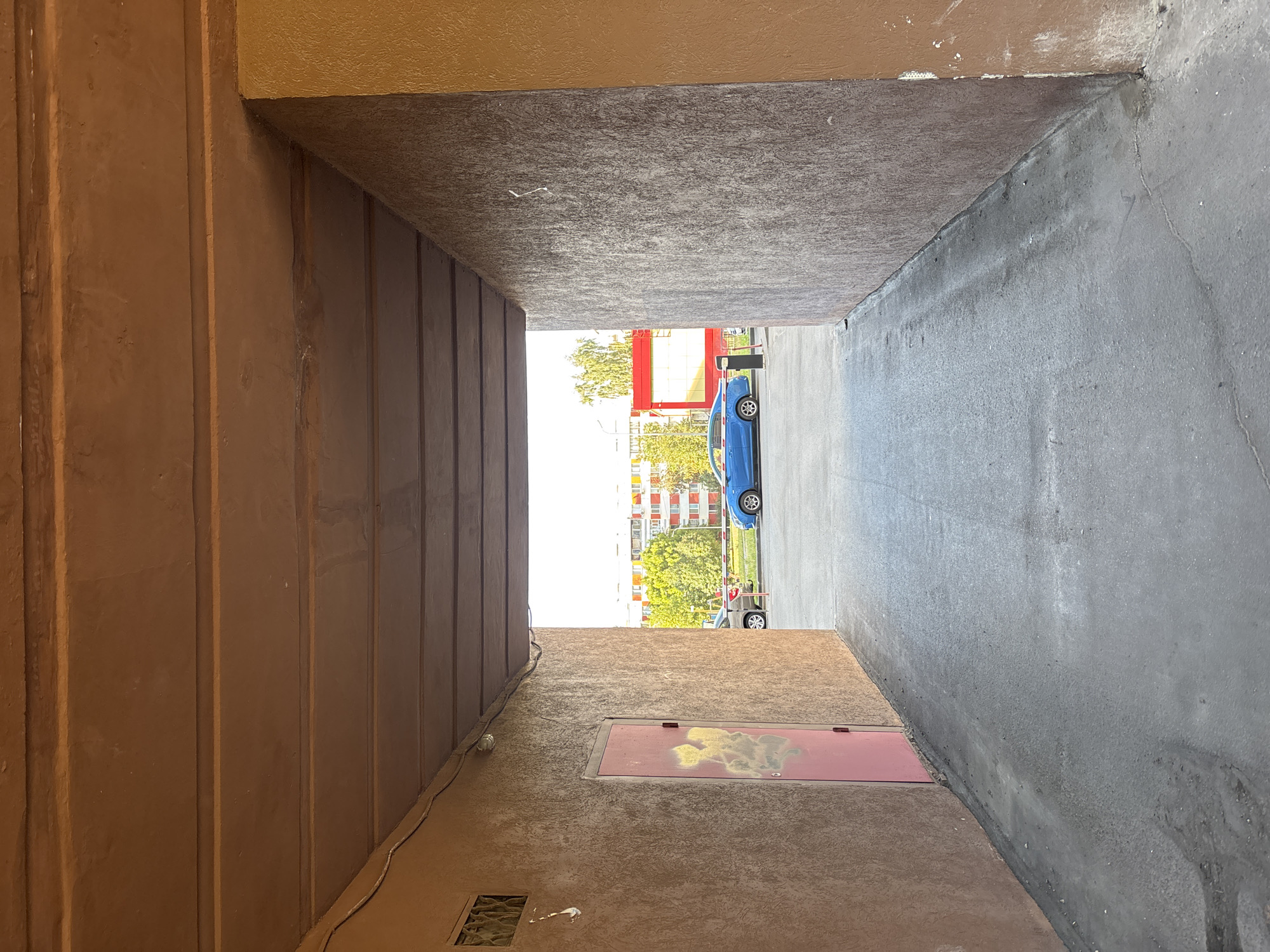 The image shows a view through a covered walkway or tunnel with a textured brown ceiling and rough, textured brown and gray walls. The walkway leads to an outdoor area with an asphalt ground. On the left wall, there is a pink door with some yellow graffiti. On the right wall, there is some visible white paint. In the background, a blue car is parked in front of a building with red and white detailing. Trees and other buildings are visible in the distance. A red barrier is also present in the background. The lighting suggests it is daytime with bright outdoor light. The location is identified as Naberezhnye Chelny, Russia.