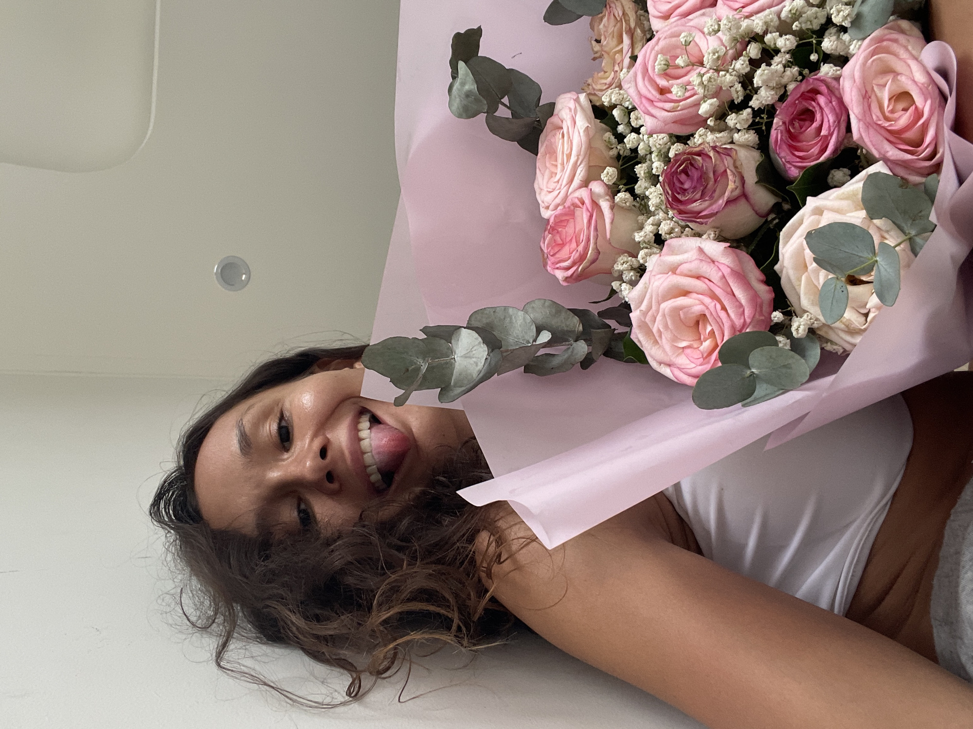 A woman with curly brown hair is lying down and holding a bouquet of pink roses and baby's breath. She is wearing a white top. The background is a plain white wall and ceiling. The image does not contain any information that would indicate the location in Phu Thuy Ward, Vietnam.