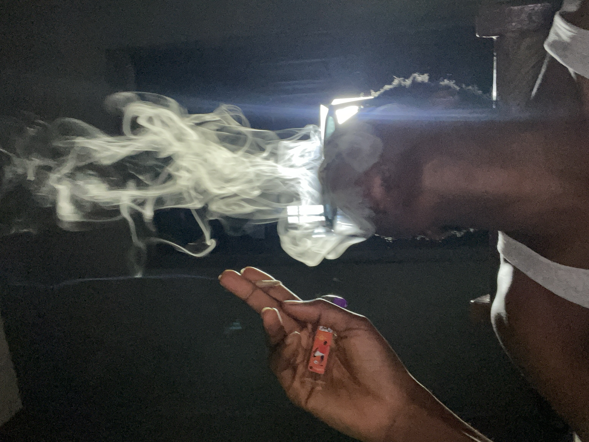 The image depicts a close-up, low-angle shot of a person exhaling a large plume of smoke. The smoke billows upwards and drifts to the right, creating ethereal, swirling patterns against a dark, out-of-focus background. The person's face is partially obscured by the smoke, but their mouth and chin are visible, and they appear to be looking upwards.
In the foreground, a dark-skinned hand is raised, with fingers extended. Clutched between the thumb and forefinger is a small, rectangular object. The object is orange with a cartoon-like depiction of a character's face and a red border. A purple ring or bead is visible on the person's ring finger. The lighting in the scene is dramatic, with a strong light source from behind and to the right, casting highlights on the hand and illuminating the smoke.
The environment appears to be indoors, with a dark wall or curtain in the background. There are hints of light coming from what could be a window or a light source behind the smoke, creating a slightly diffused glow. The presence of the smoke suggests an activity involving smoking. The overall mood is atmospheric and somewhat mysterious due to the interplay of light and shadow. The specific location in Nkpor, Nigeria, cannot be determined from the visual information provided in the image alone.