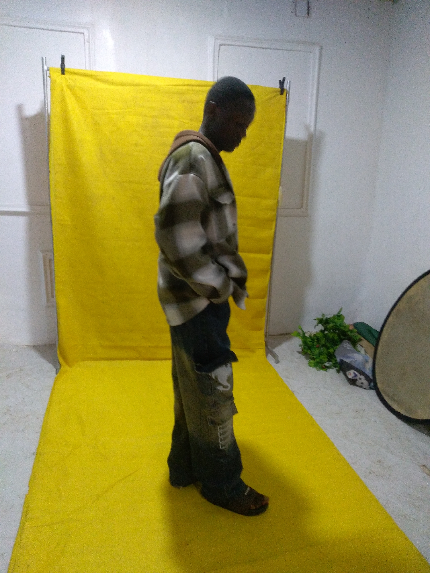 A young Black male stands in a studio, facing to the right of the frame, with his head bowed. He is wearing a long-sleeved, checkered shirt over a hooded sweatshirt, and baggy, dark-colored pants with large pockets. The backdrop is a bright yellow fabric stretched across a stand, and a matching yellow mat covers the floor. To the right of the frame, a round, reflective object, possibly a photographic reflector, leans against a white wall. A small cluster of green plants is also visible on the floor in the background. The room appears to be an indoor space, possibly a photography studio, with white walls and a tiled floor. The overall impression is one of a staged setting, possibly for a portrait or a fashion shoot. The lighting is bright and even, illuminating the subject and the yellow backdrop.