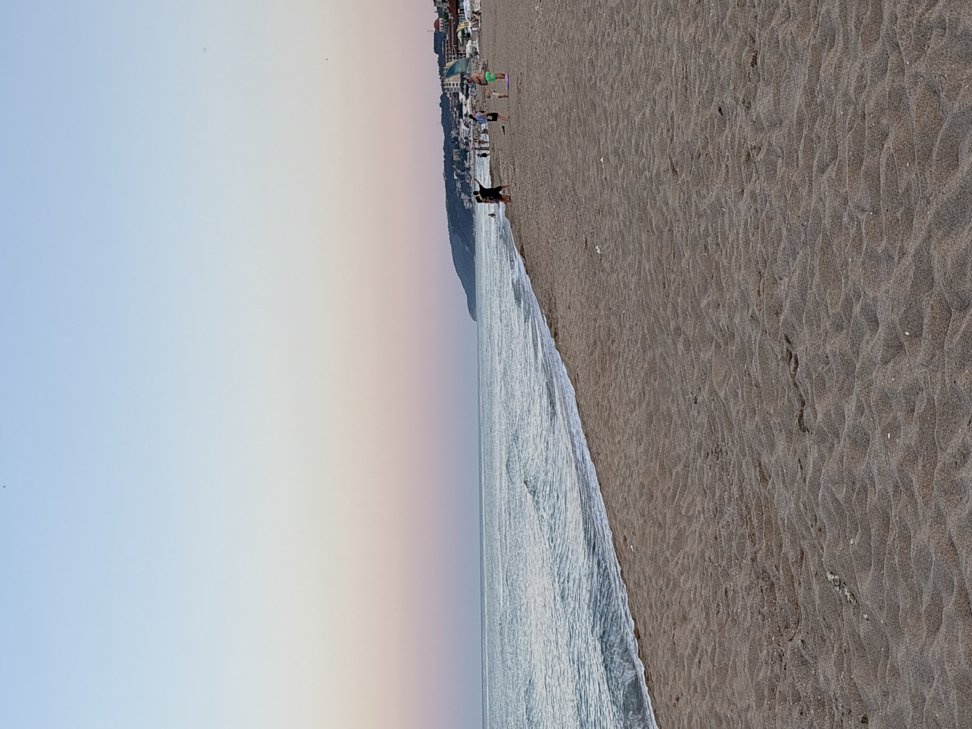 This is a photograph of a beach scene in Obzor, Bulgaria, captured at dusk. The sky transitions from a pale blue at the top to a soft pink hue near the horizon. The sea is calm with small waves lapping at the sandy shore. In the distance, a hilly coastline is visible, dotted with buildings that appear to be part of the town. Several people are present on the beach. Some are walking, while others are standing and interacting. One person in the foreground appears to be pointing towards the sea. The sand is textured with ripples, indicative of wind or receding waves. The overall impression is a peaceful evening by the sea.